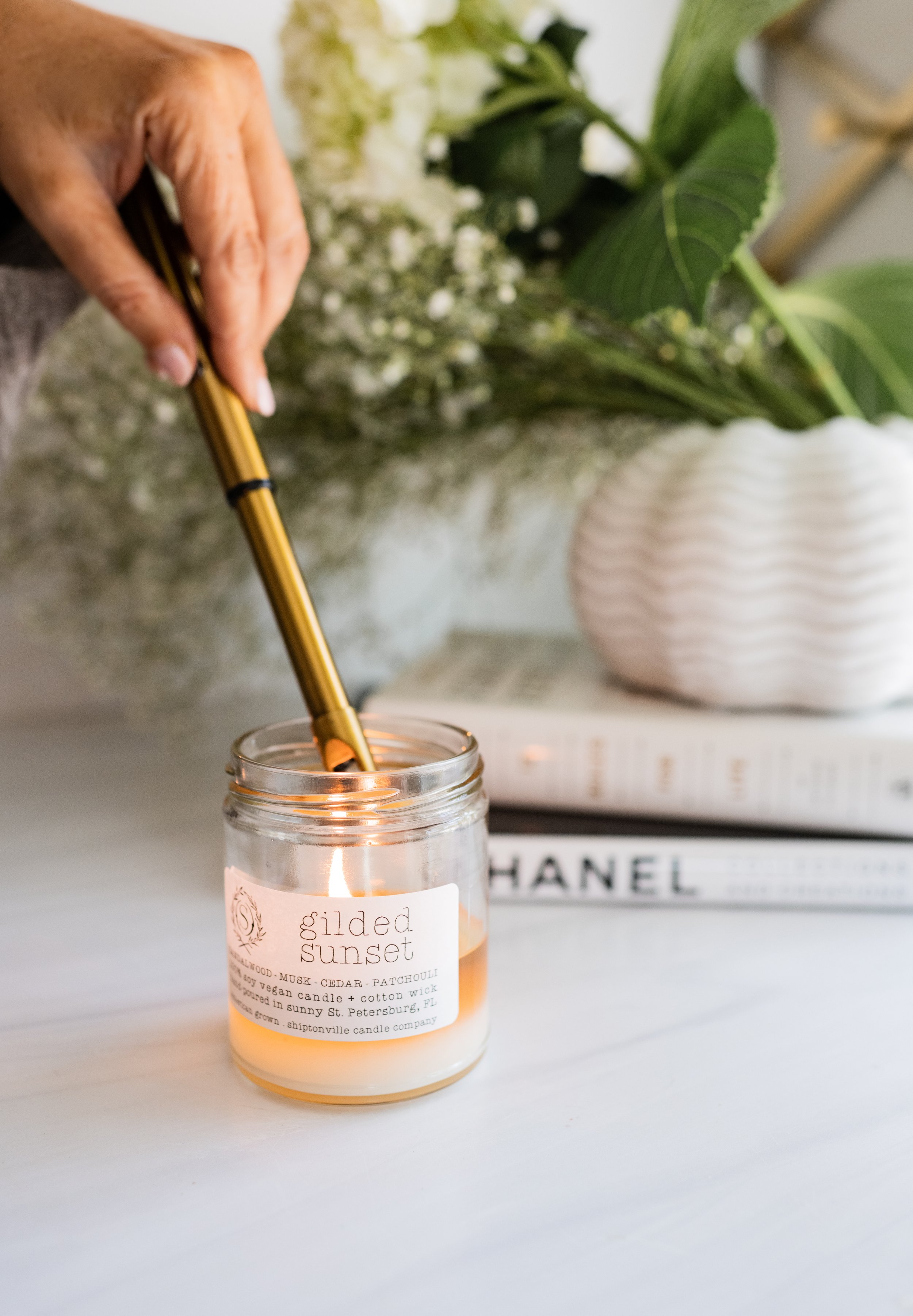 Person lighting a candle labeled 'gilded sunset' with a gold lighter, with green plants and a stack of books in the background.