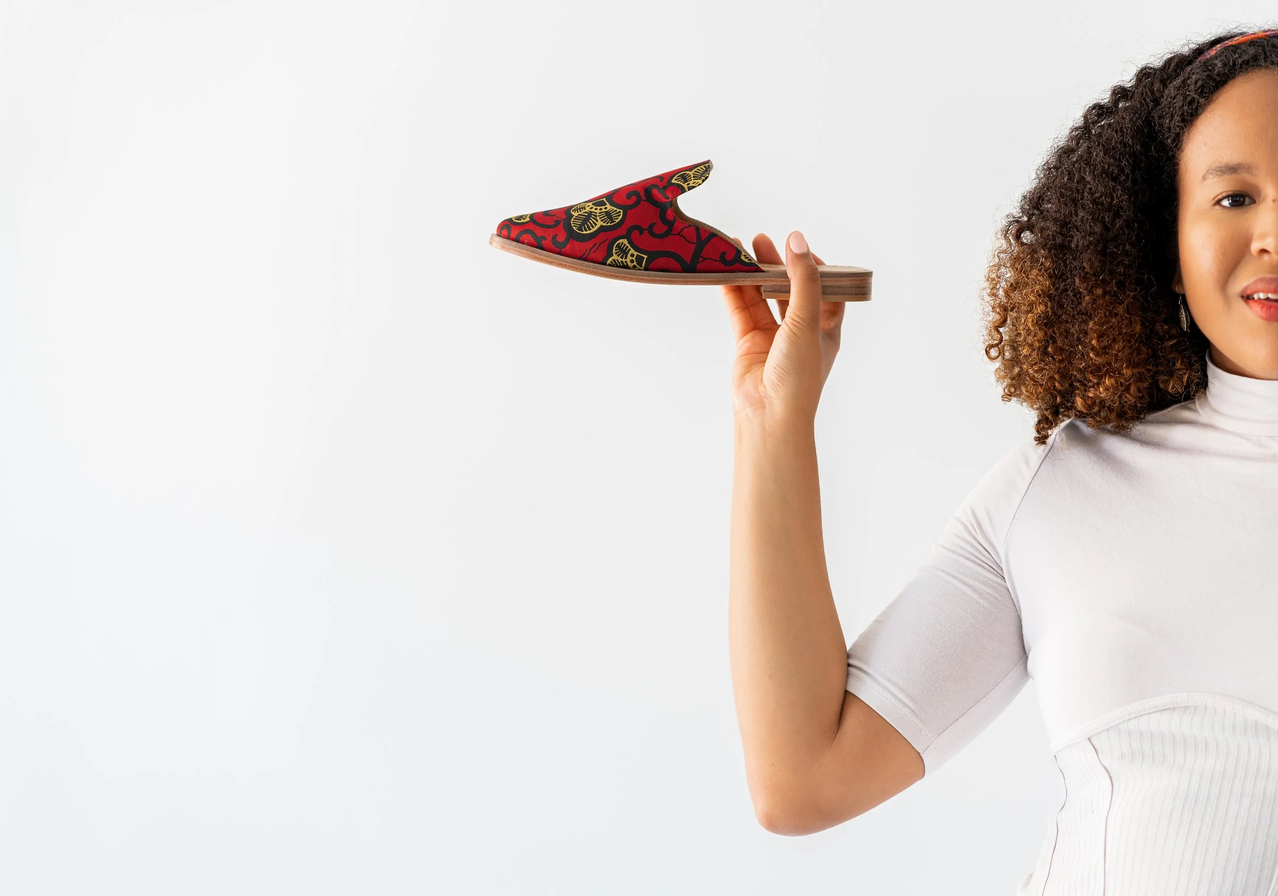 A woman with curly hair holding up a red and black patterned slip-on shoe against a plain white background.