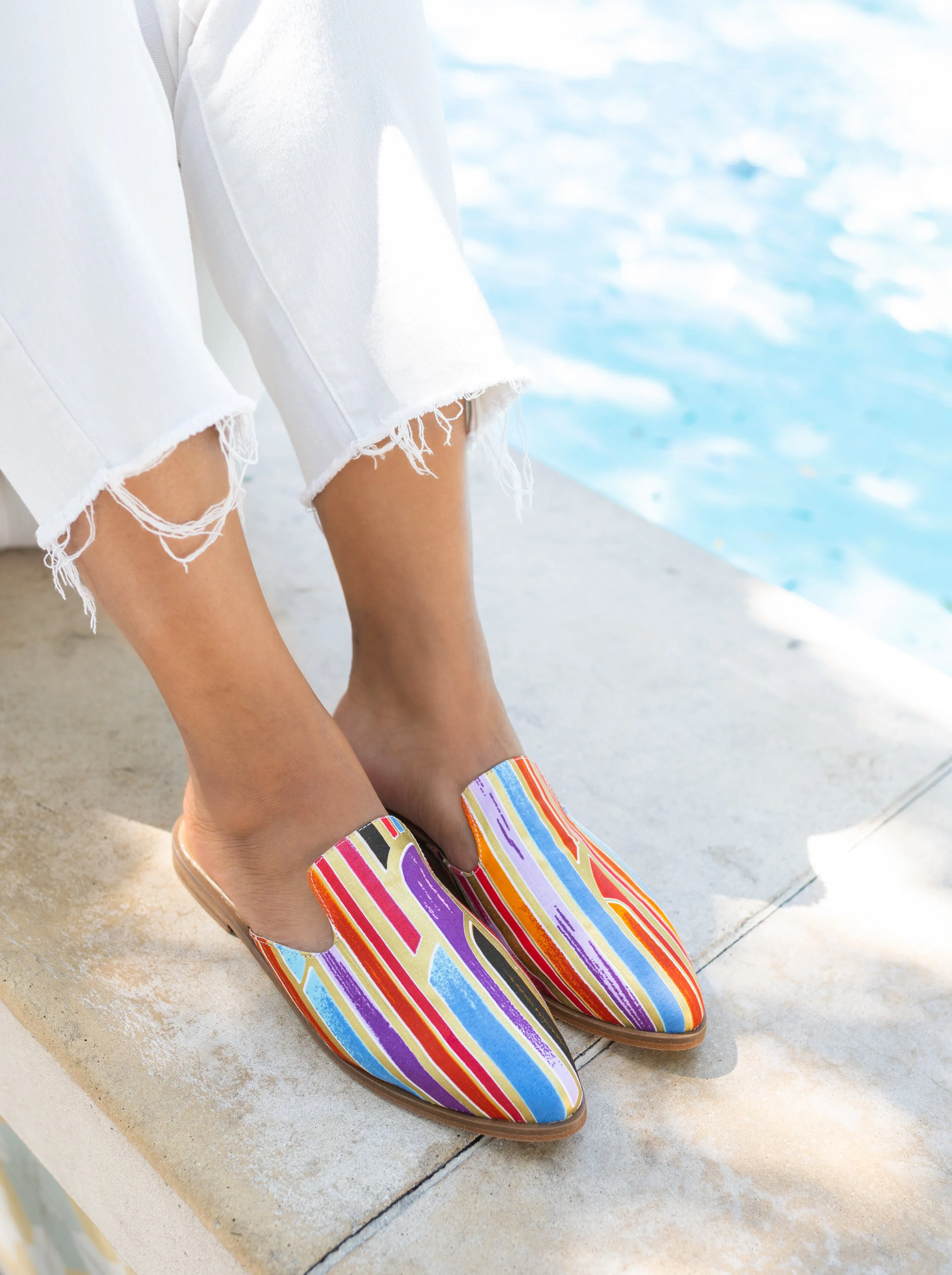 Person wearing colorful striped slip-on shoes sitting by a pool with white ripped jeans.