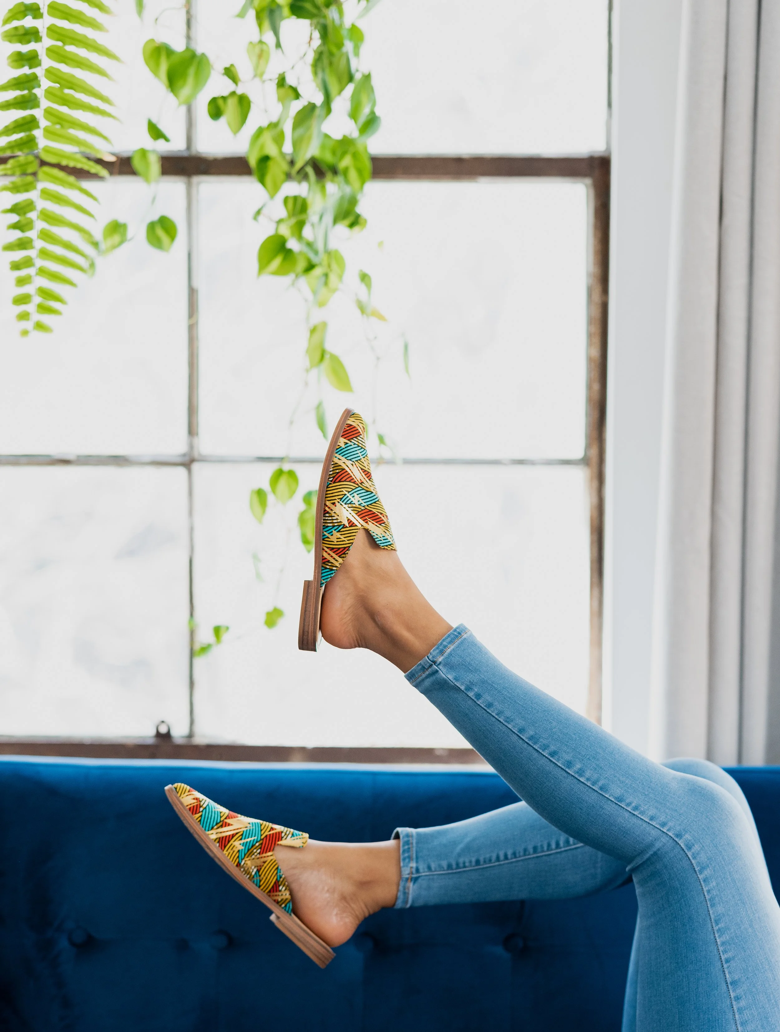 Person wearing colorful patterned shoes with legs raised, sitting on a blue sofa in front of a large window with greenery outside.