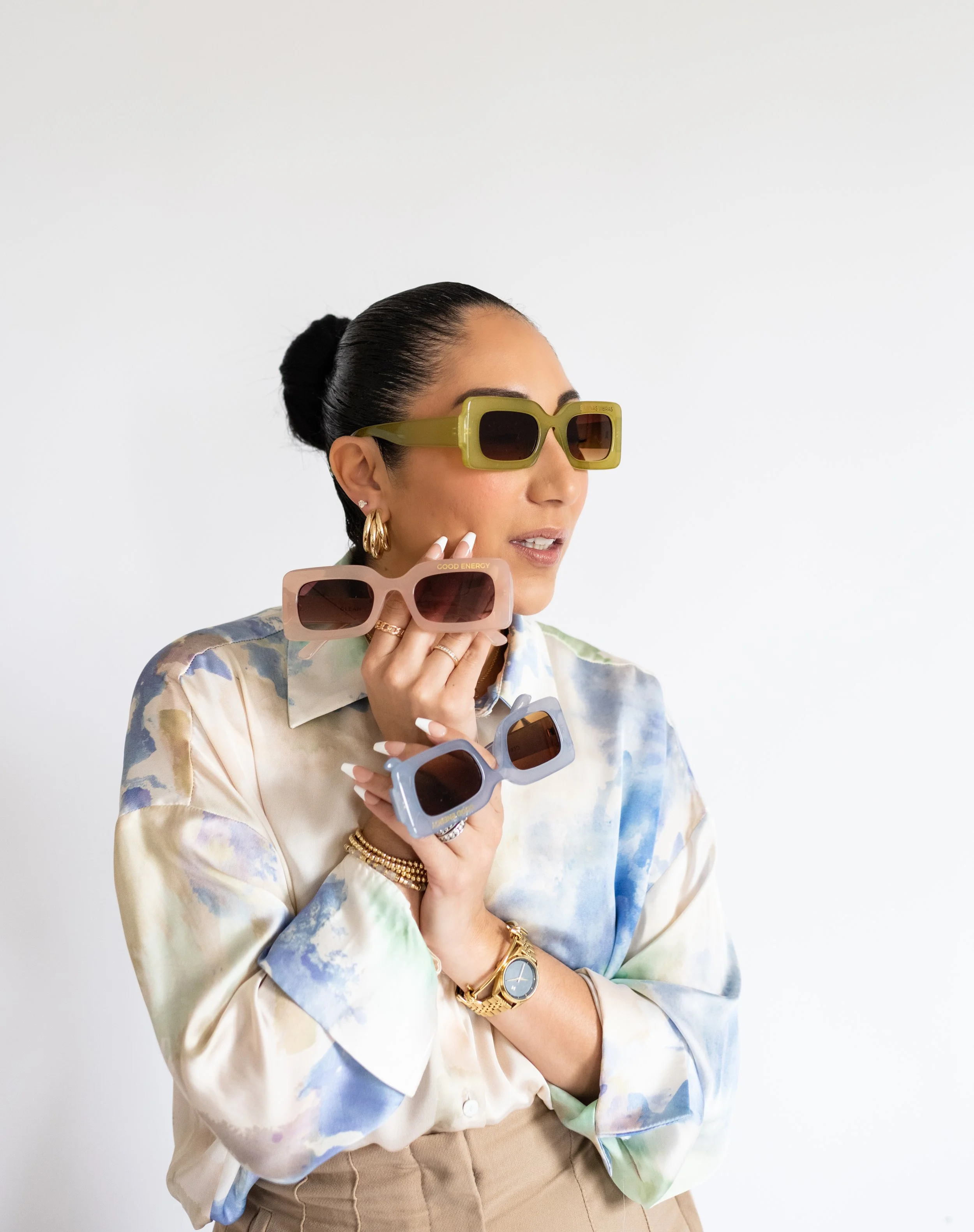 A woman holding colorful sunglasses, wearing a floral blouse, gold jewelry, and sunglasses, against a plain white background.