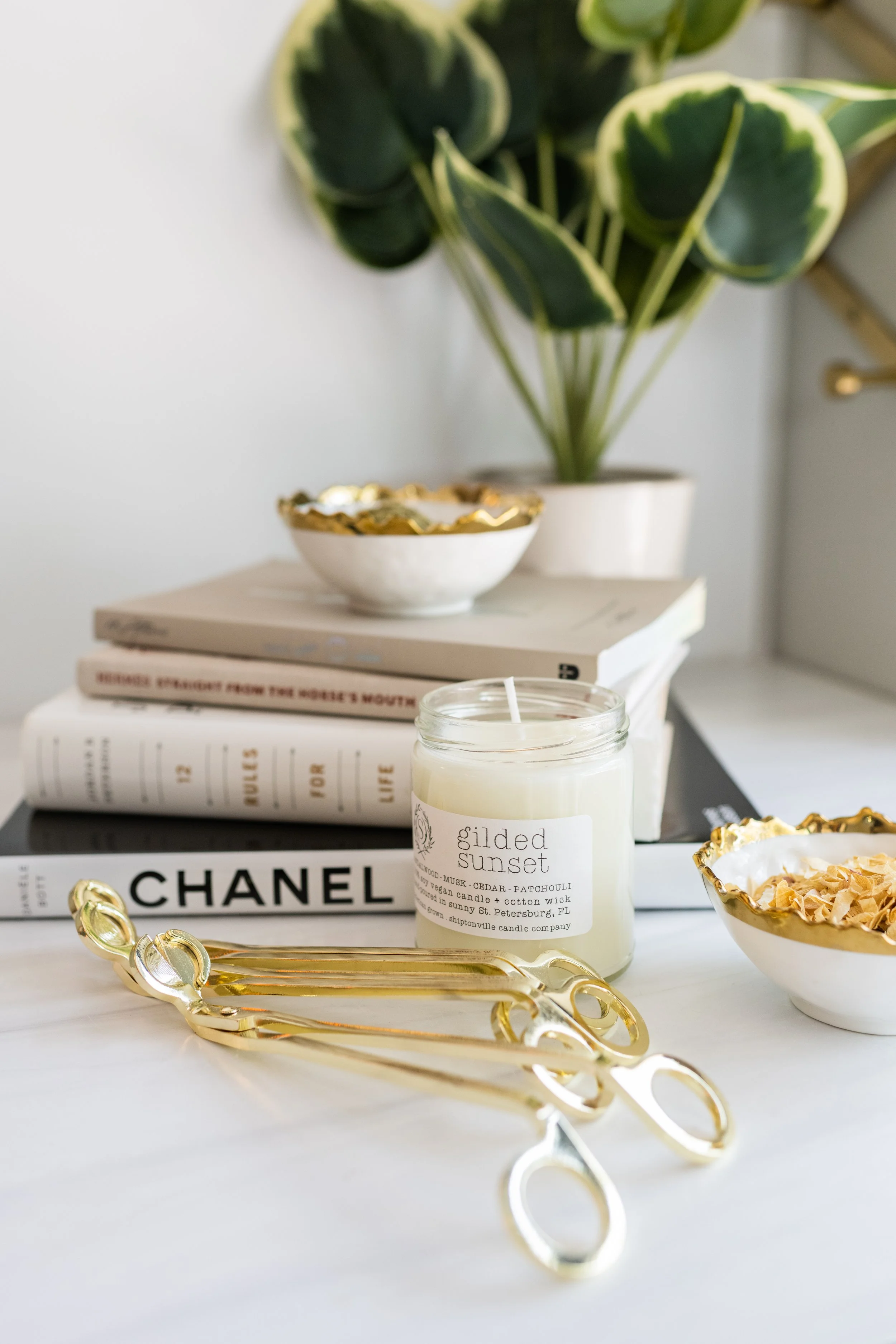 A collection of decorative items on a white surface, including a jar candle labeled "Gilded Sunset," a pair of gold scissors, a bowl filled with potpourri, a stack of books, and a green potted plant in the background.