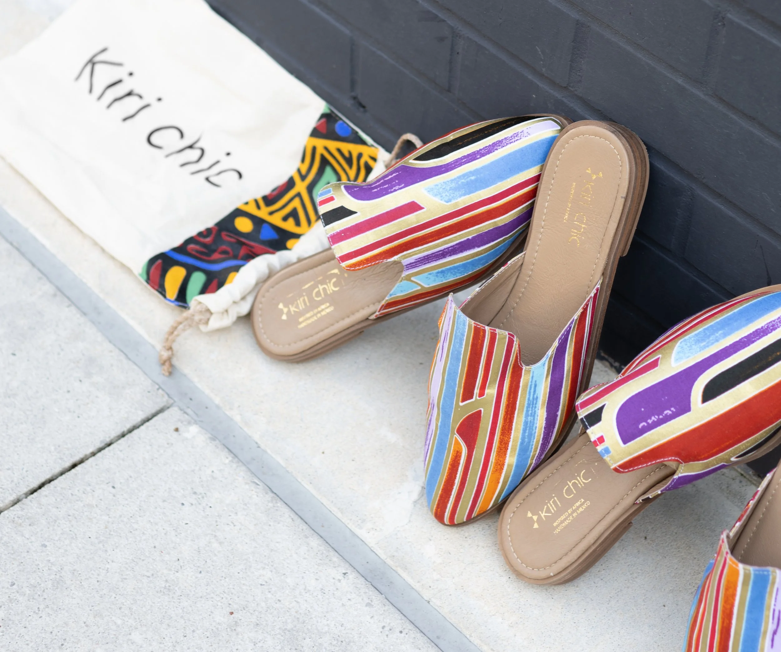 Pair of colorful striped slip-on sandals, and a cloth bag with the name 'Kiri Chic' written on it, placed against a black wall and on a concrete sidewalk.