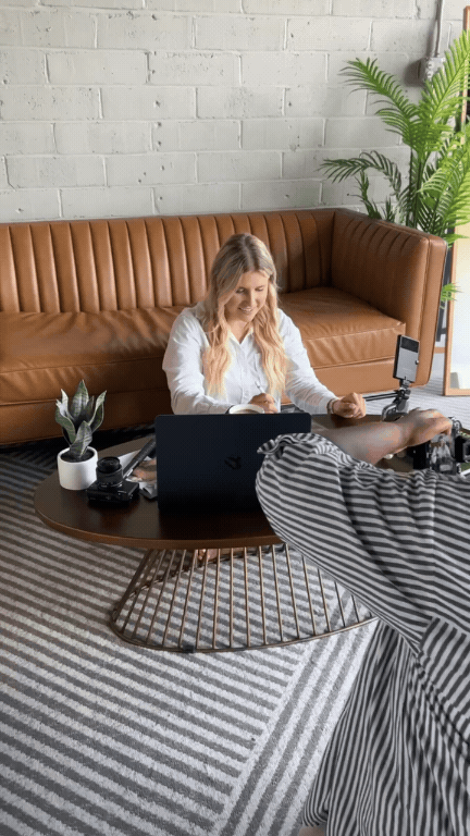 A woman sitting on the floor using her phone during a video shoot, with a cameraman filming her, in a modern living room with a brown leather couch, potted plants, a round coffee table, and a striped rug.