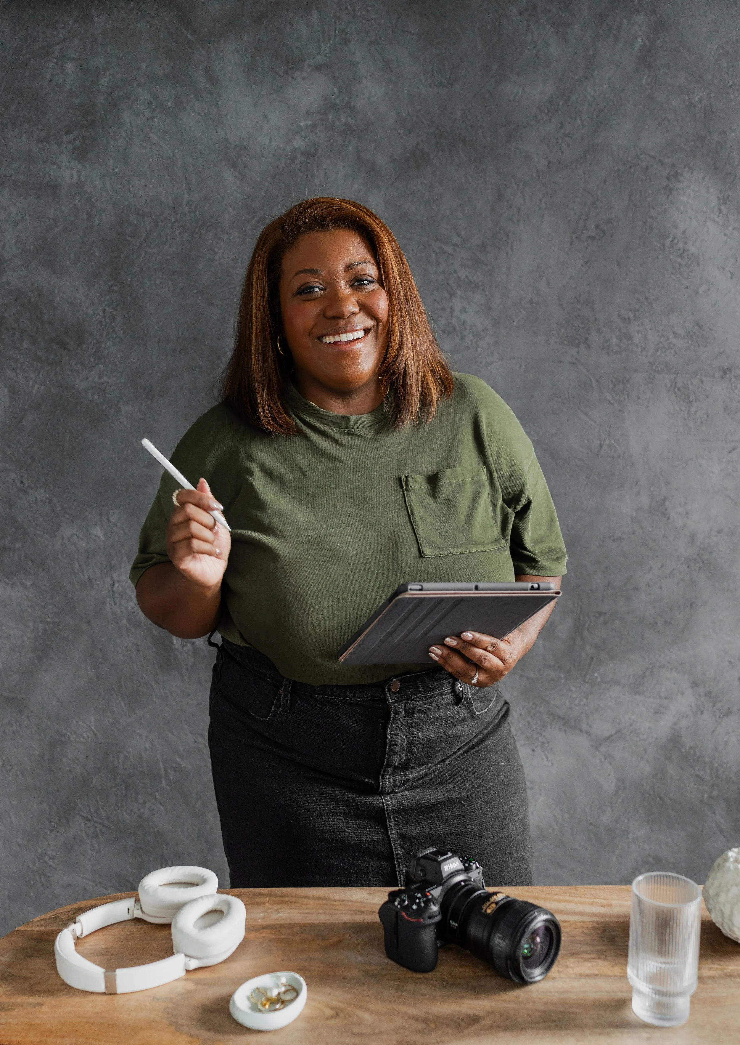 A smiling woman with shoulder-length brown hair holding a stylus and a tablet, standing behind a wooden table with a Nikon camera, white headphones, a glass of water, and jewelry on it, against a textured gray wall.