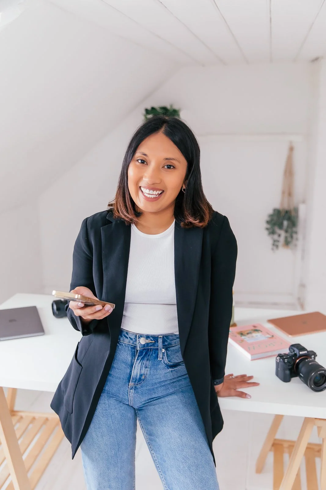 Smiling woman in black blazer and jeans holding a smartphone, standing in a bright, minimalistic room with a white table, books, and a camera.