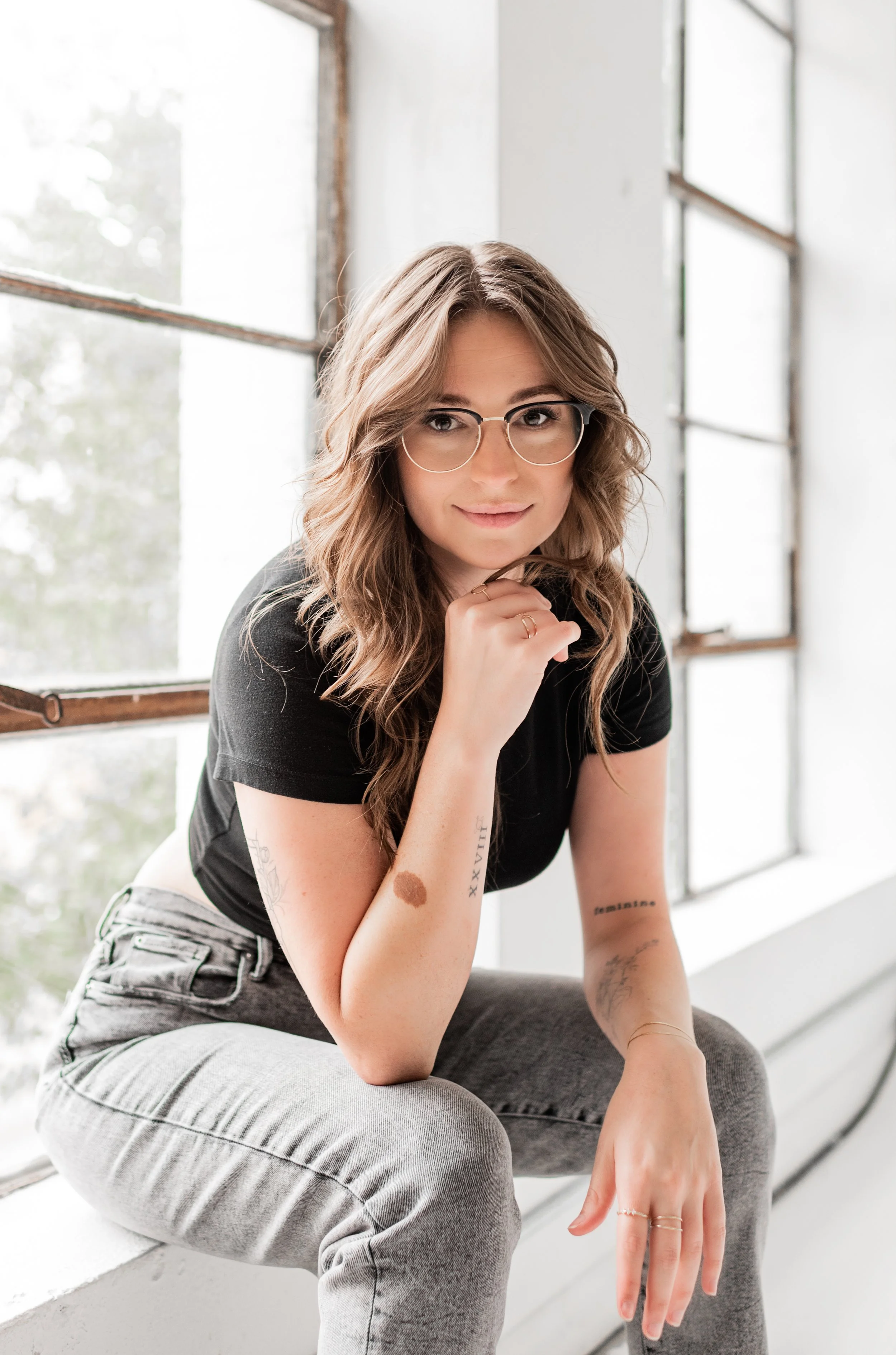 A young woman with wavy hair, glasses, and tattoos on her arm, sitting on a windowsill indoors, looking into the camera with a slight smile.