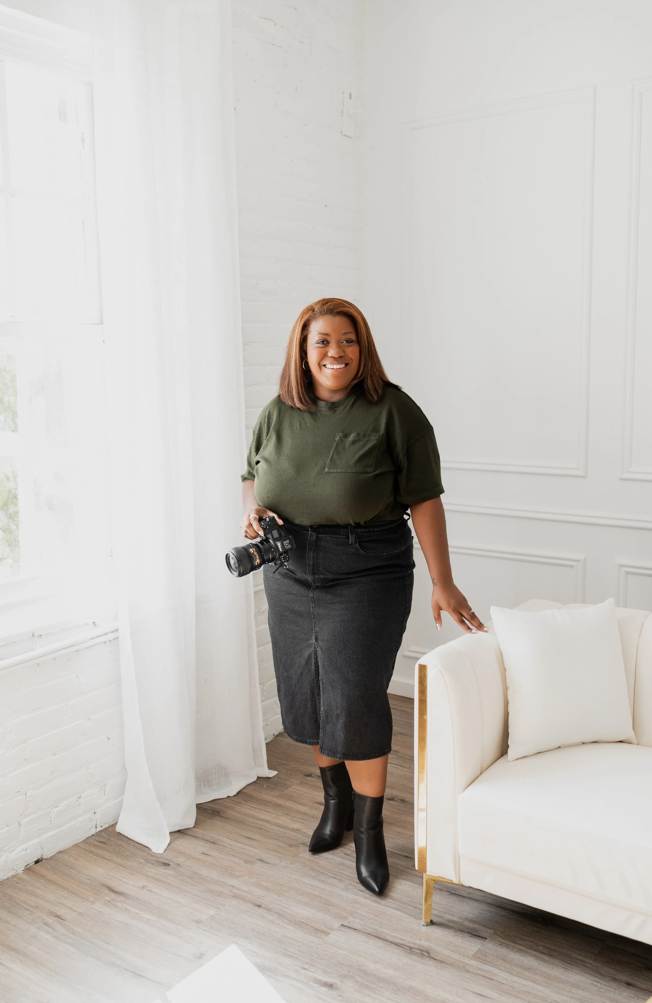 A woman with shoulder-length hair, wearing a green t-shirt, black skirt, and black boots, is standing in a bright room with white walls and curtains, holding a camera.