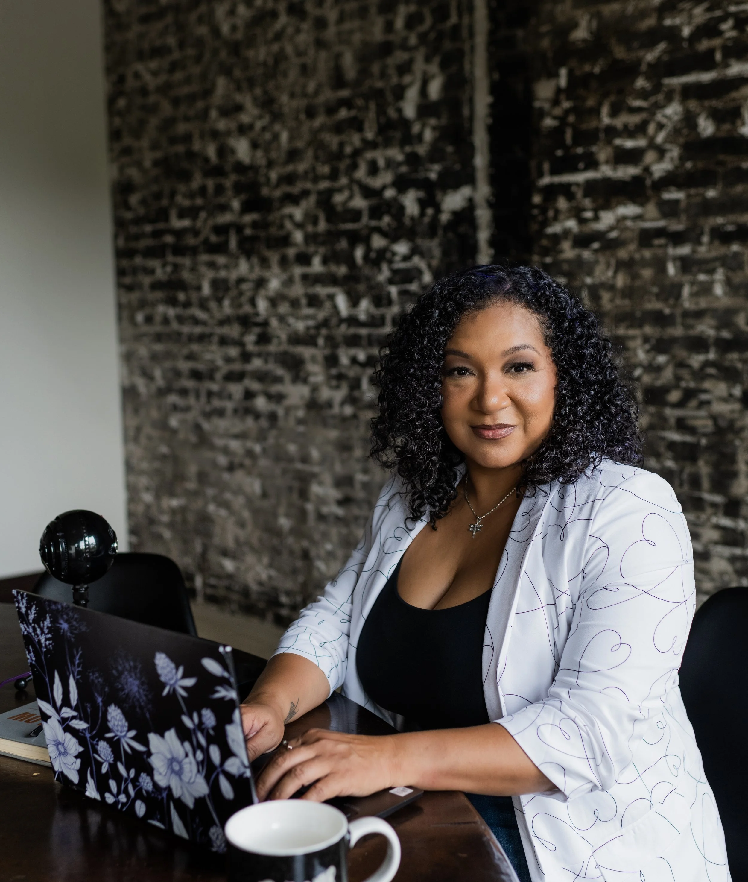 A woman with curly black hair sitting at a desk with a laptop, a mug, and a notebook, in front of a brick wall.