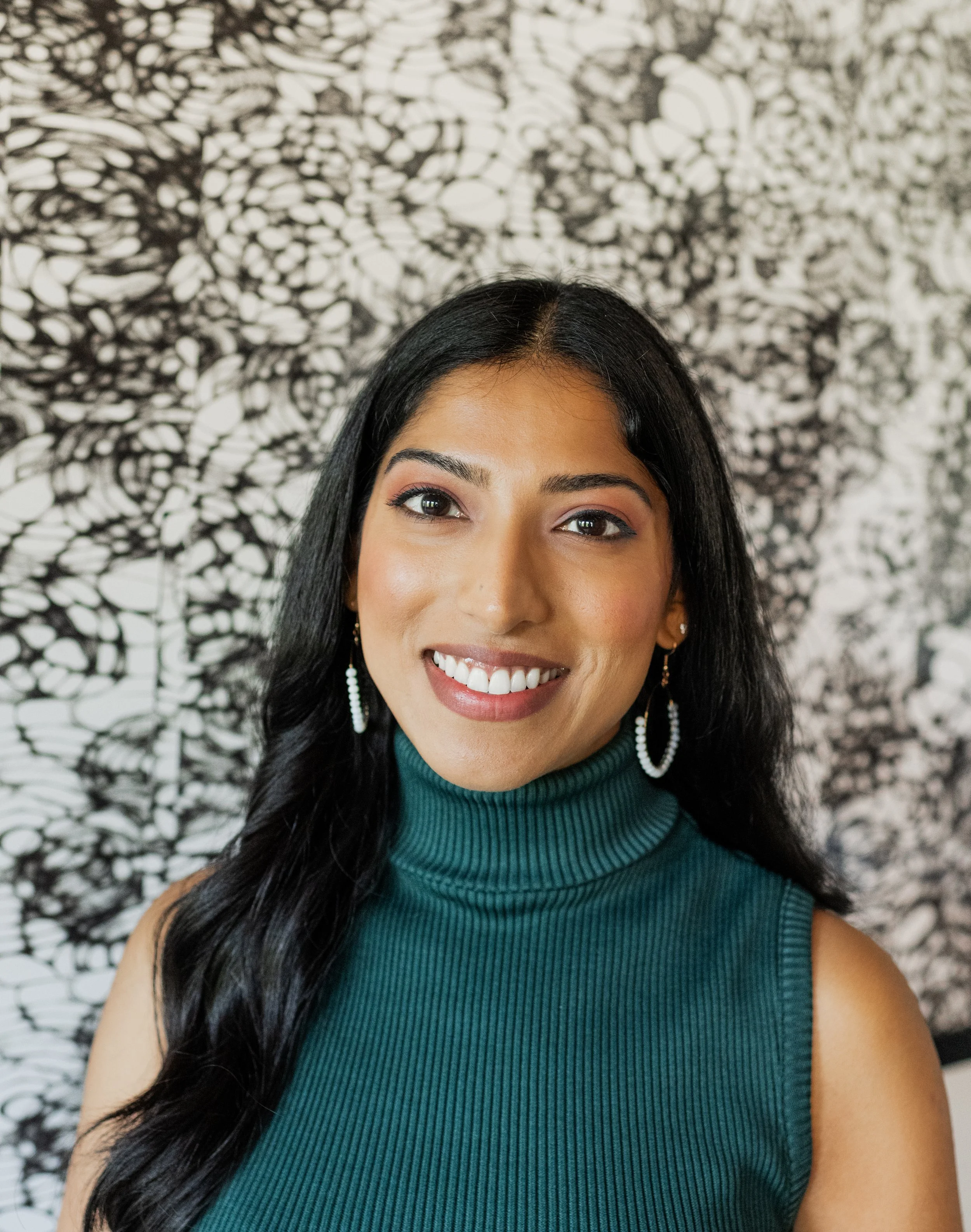 A young woman with long dark hair, wearing a teal sleeveless turtleneck top and hoop earrings, smiling in front of a black and white abstract art background.