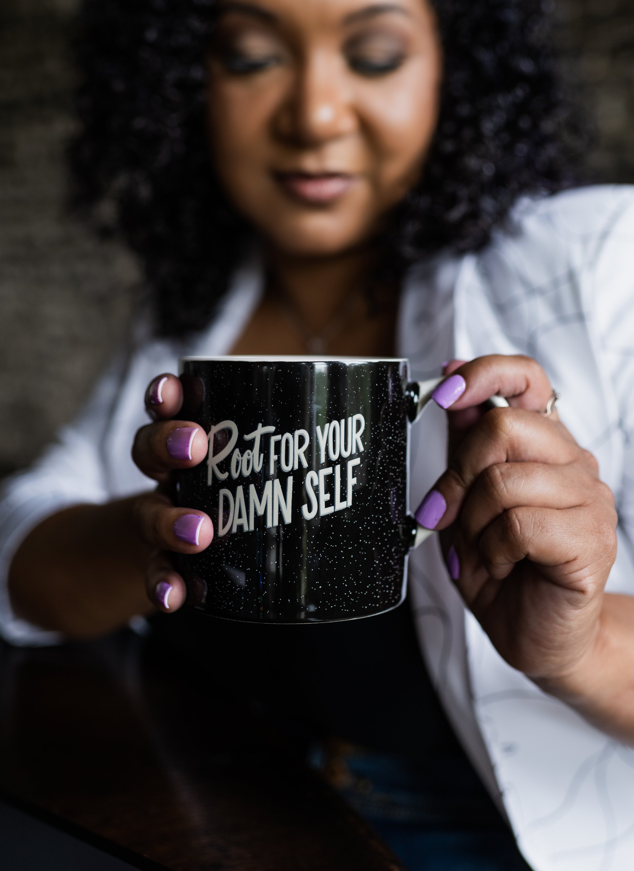 A woman with curly hair and purple-painted nails holding a black mug that says 'Root for your damn self' in white text.