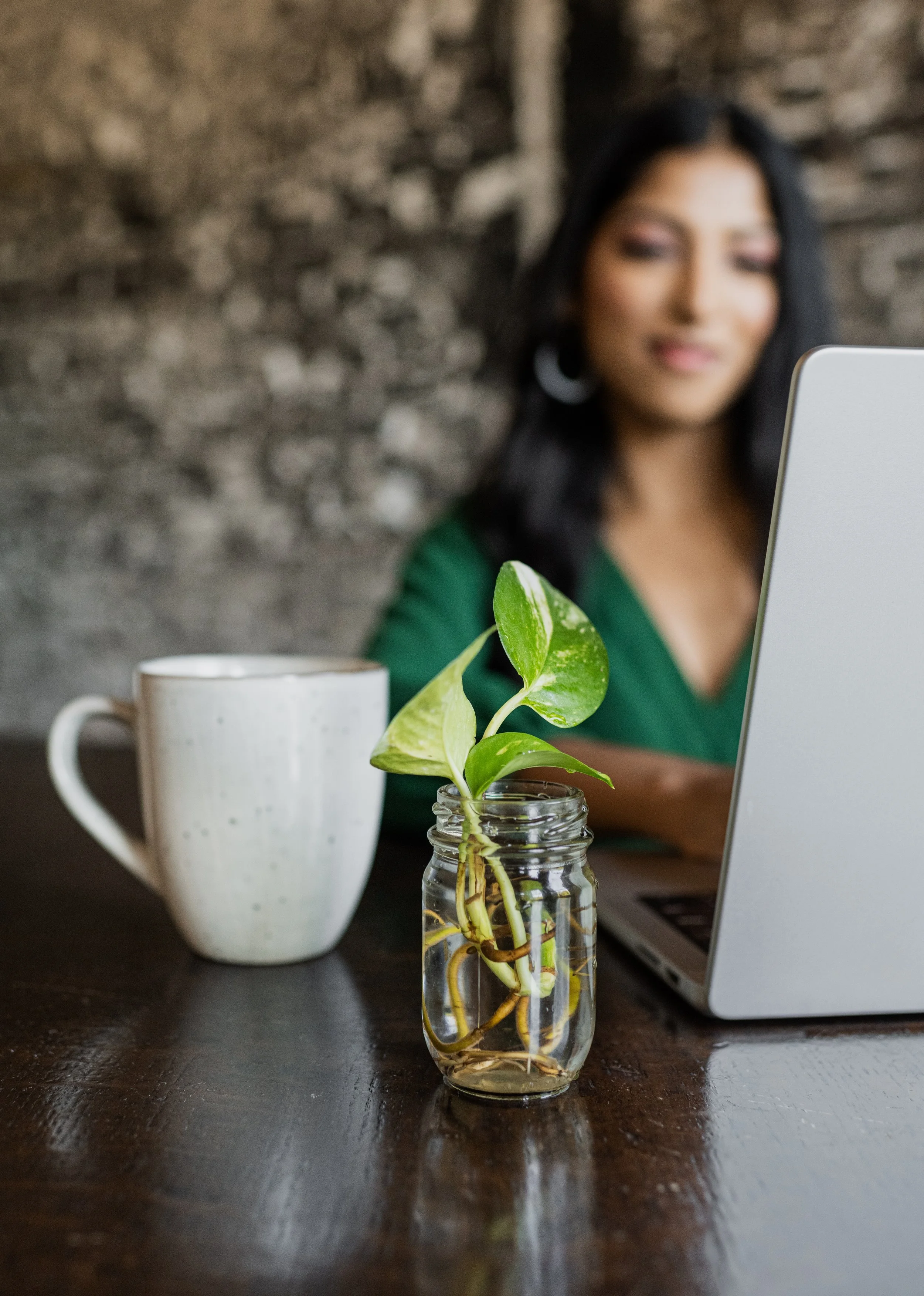 A woman sitting at a table with a jar of green houseplant and a white mug, working on a laptop.