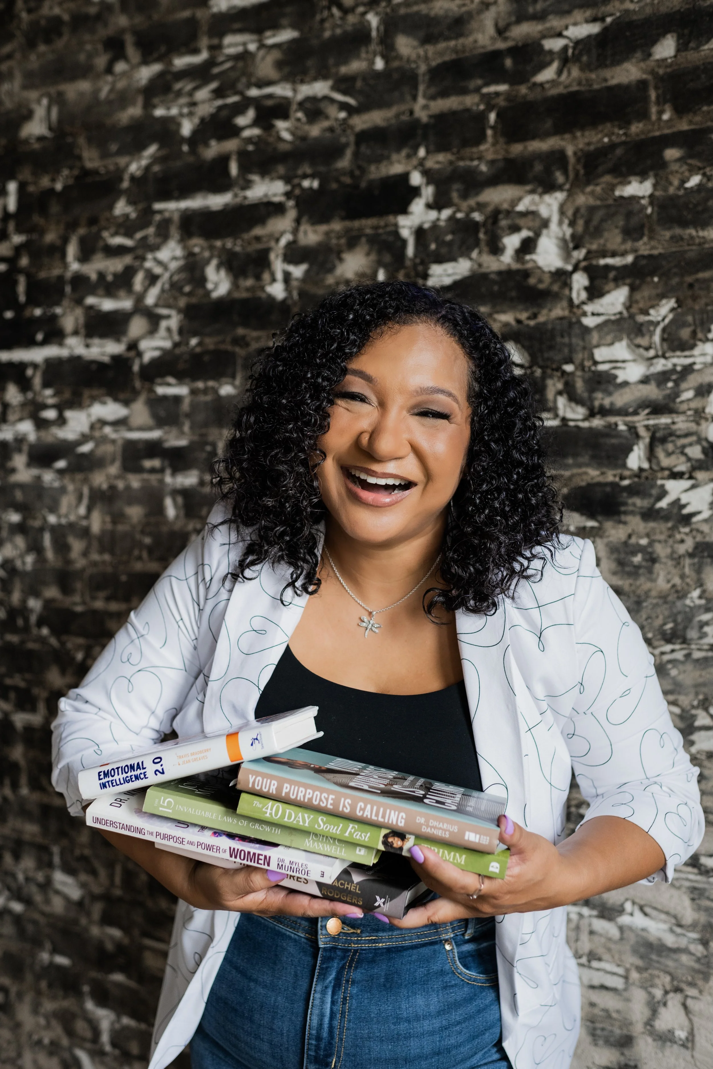 Happy woman holding a stack of five books against a brick wall.
