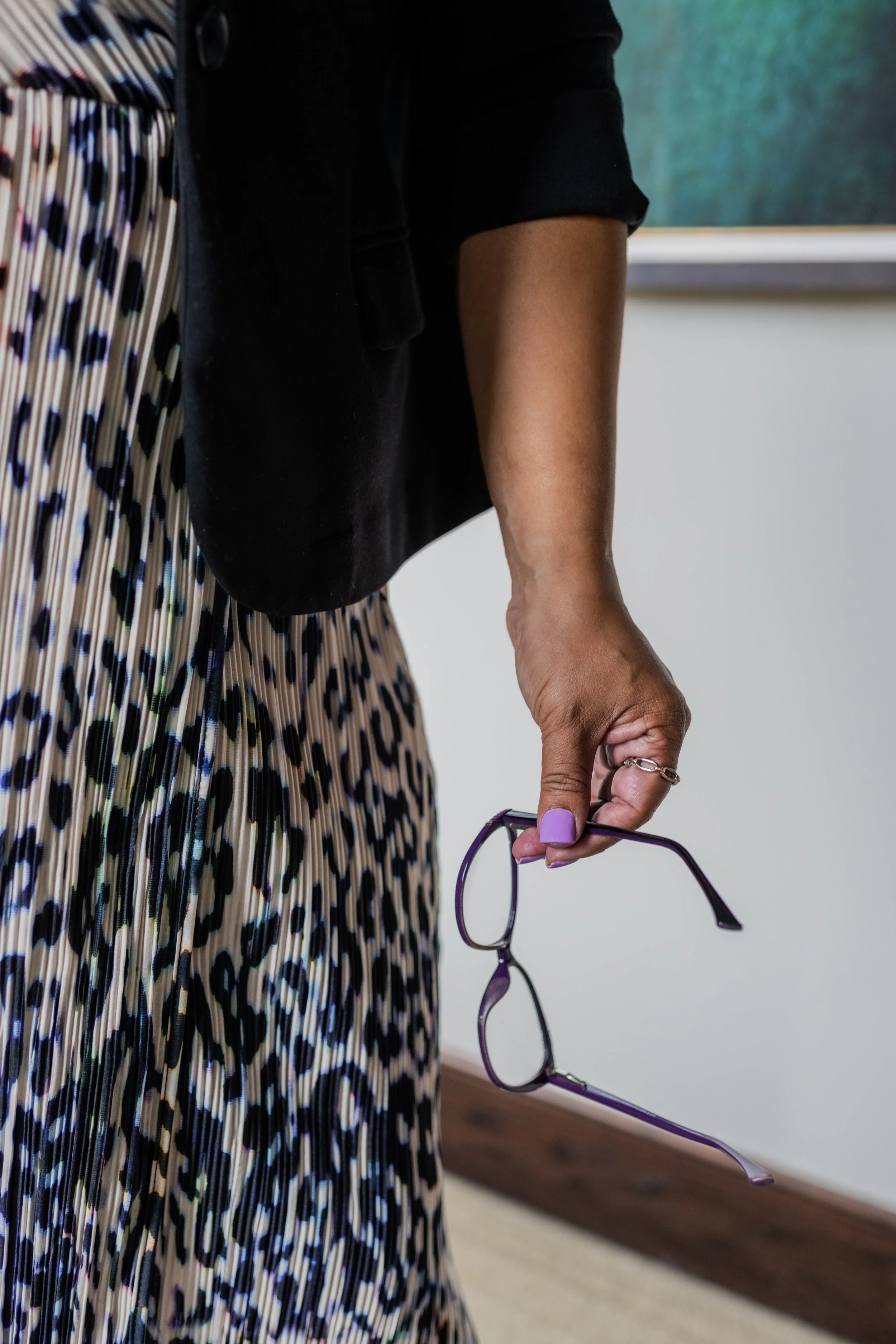 A person wearing a black blazer, leopard print pleated skirt, and purple nail polish, holding a pair of purple glasses in their right hand.