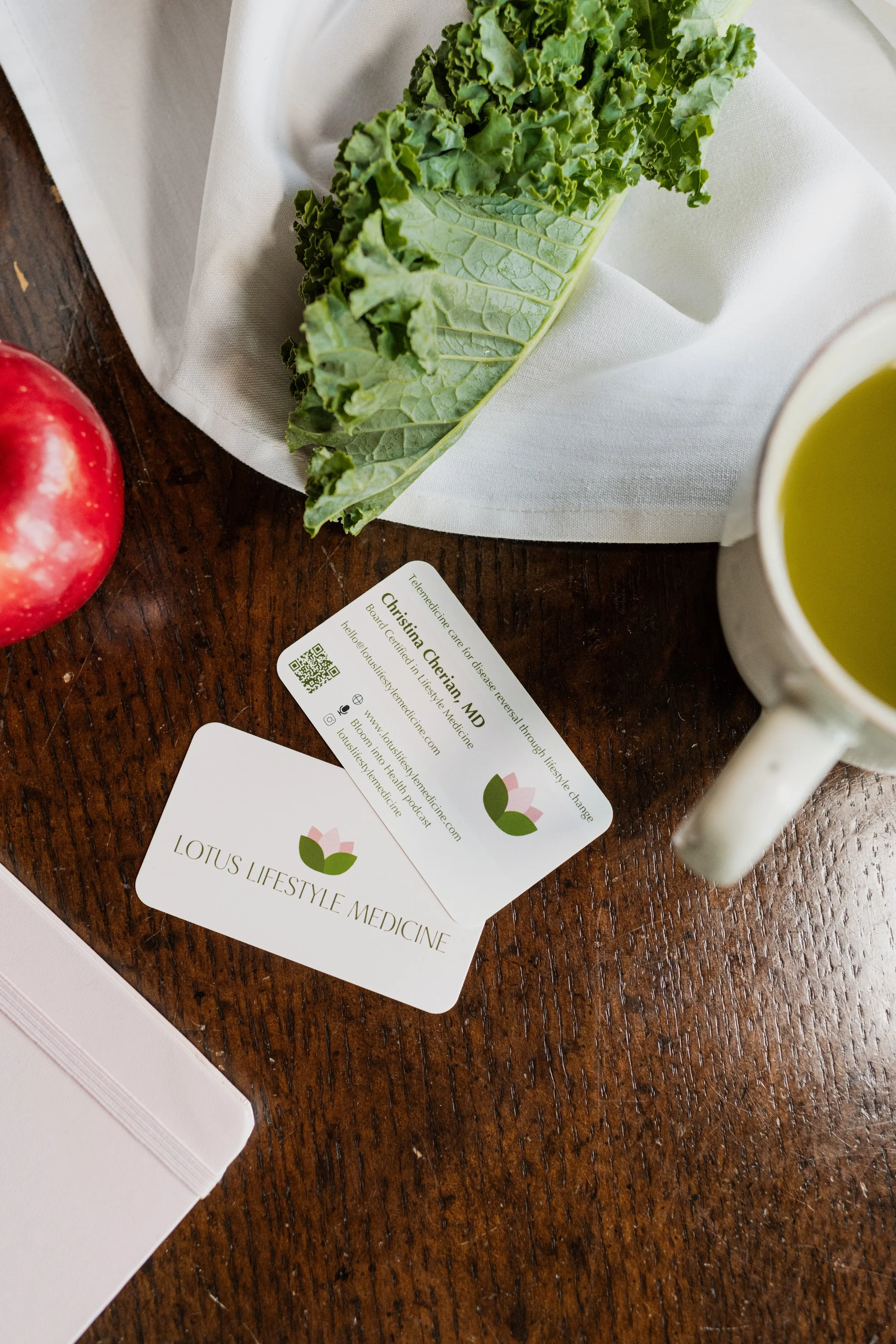 A wooden table with a business card for Lotus Lifestyle Medicine, a cup of green tea, a fresh apple, a large kale leaf, and a white notebook.
