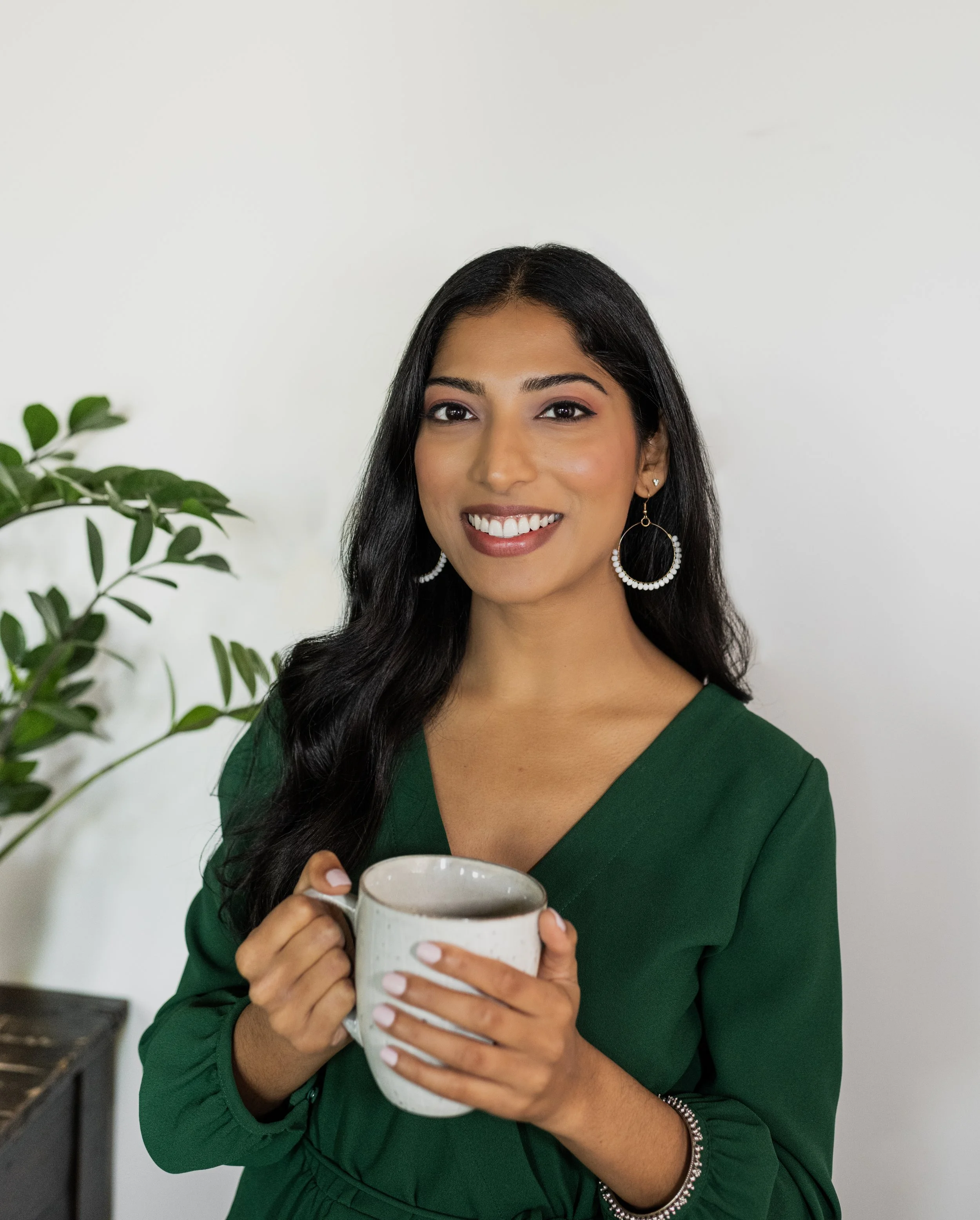 A woman with long dark hair wearing a green dress and hoop earrings holding a white mug, smiling in front of a white wall and a plant.