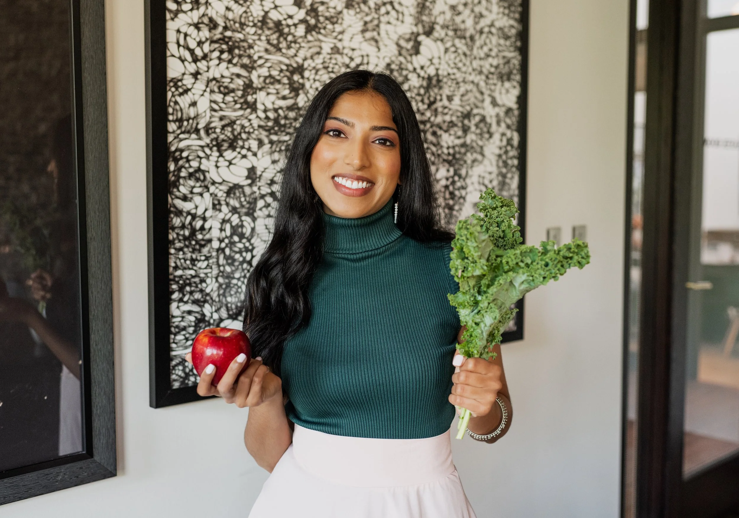 A woman with long black hair smiling, holding an apple in her right hand and a bunch of leafy greens in her left, standing indoors in front of abstract black and white artwork.