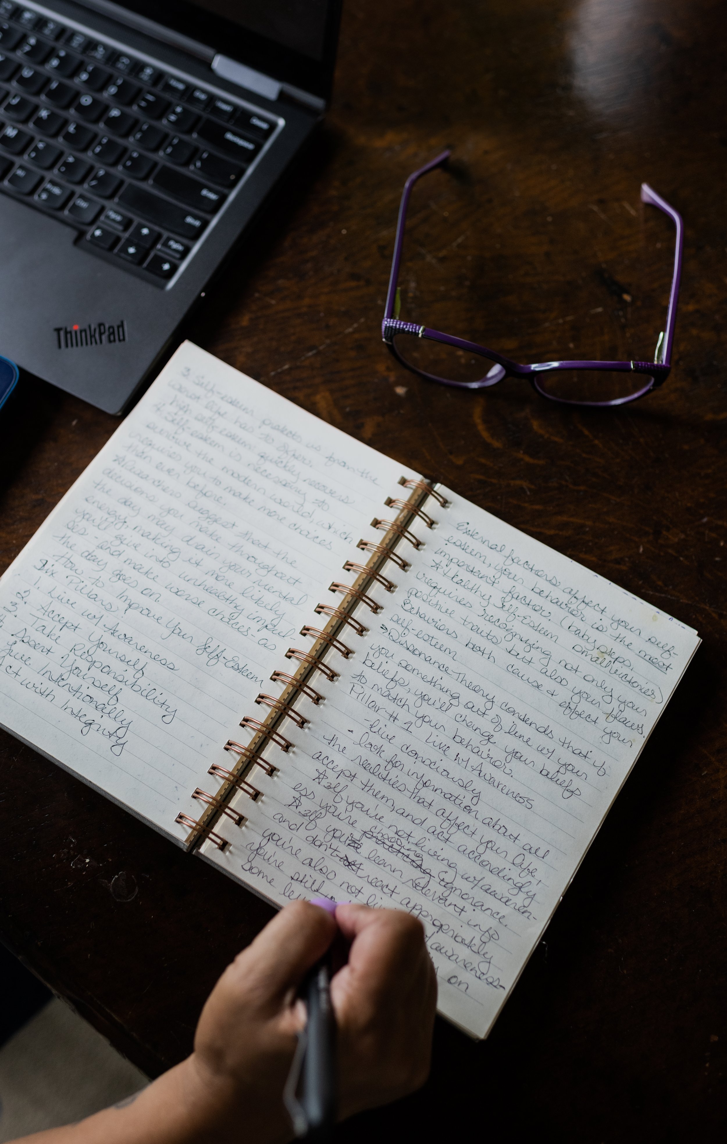 A person with purple nails taking notes in a spiral notebook on a wooden desk, with purple glasses, a black Lenovo ThinkPad laptop, and a blue phone nearby.