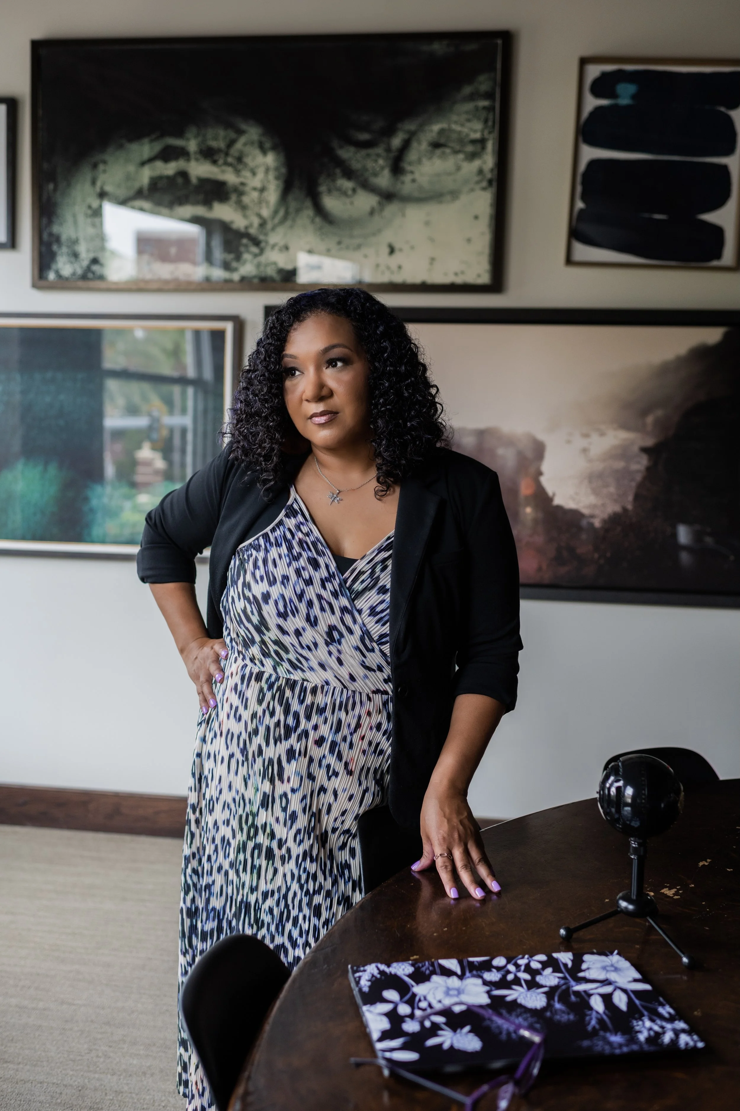A woman with curly black hair standing in an art gallery, wearing a leopard print dress and black blazer, with abstract paintings on the wall behind her.