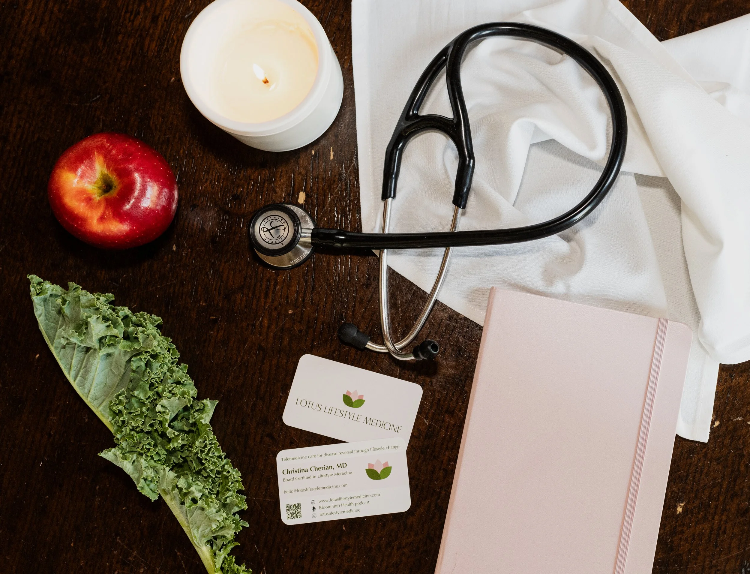 A stethoscope, an apple, a candle, a romaine lettuce leaf, a pink notebook, and a business card for Lotus Lifestyle Medicine on a wooden table.