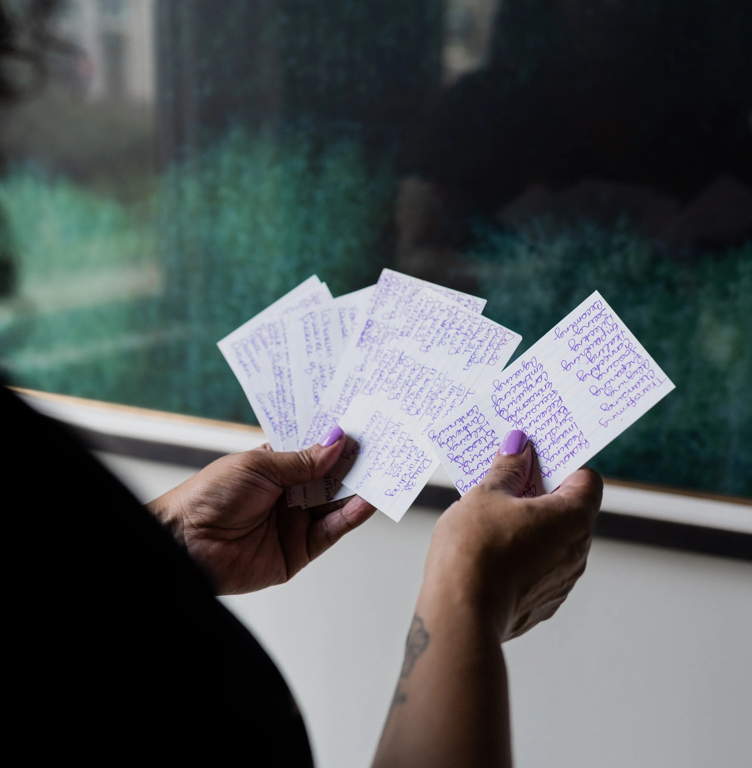 A person holding several handwritten notes with purple ink, reading them near a large window with a green landscape outside.