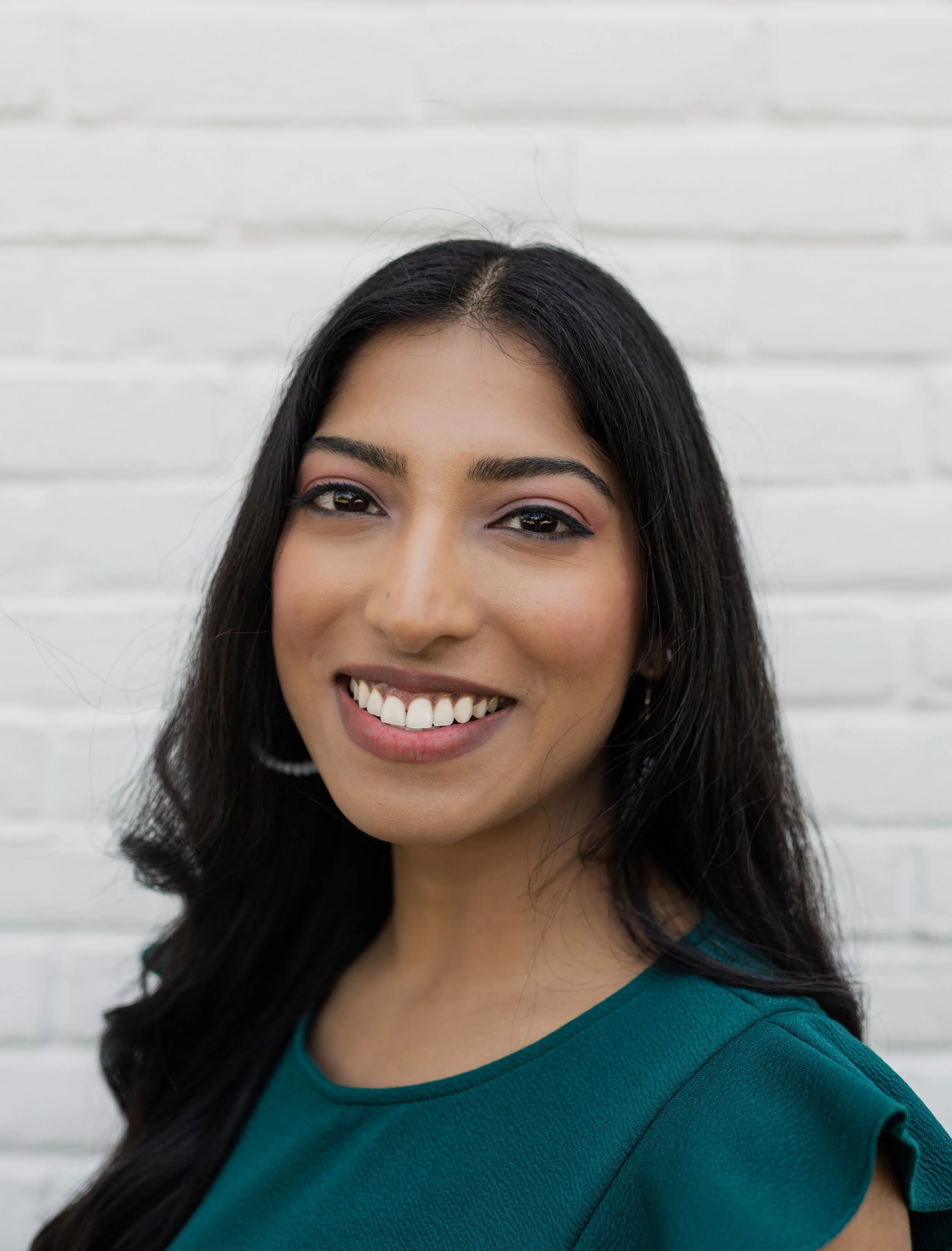 A woman with long dark hair, smiling, wearing a teal top, standing against a white brick wall.