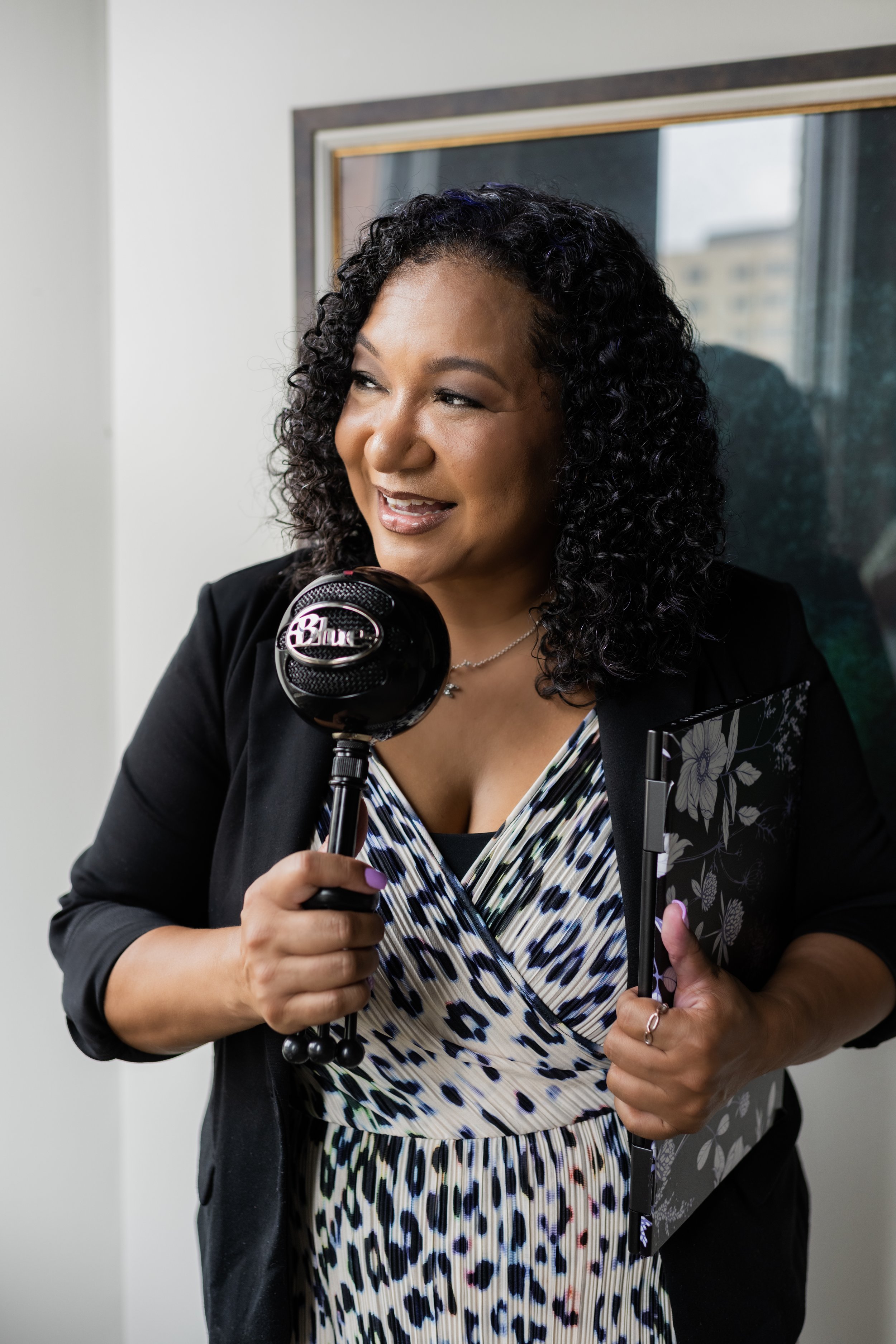 A woman with curly black hair holding a black microphone and a clipboard, smiling and looking to the side, in an indoor setting.