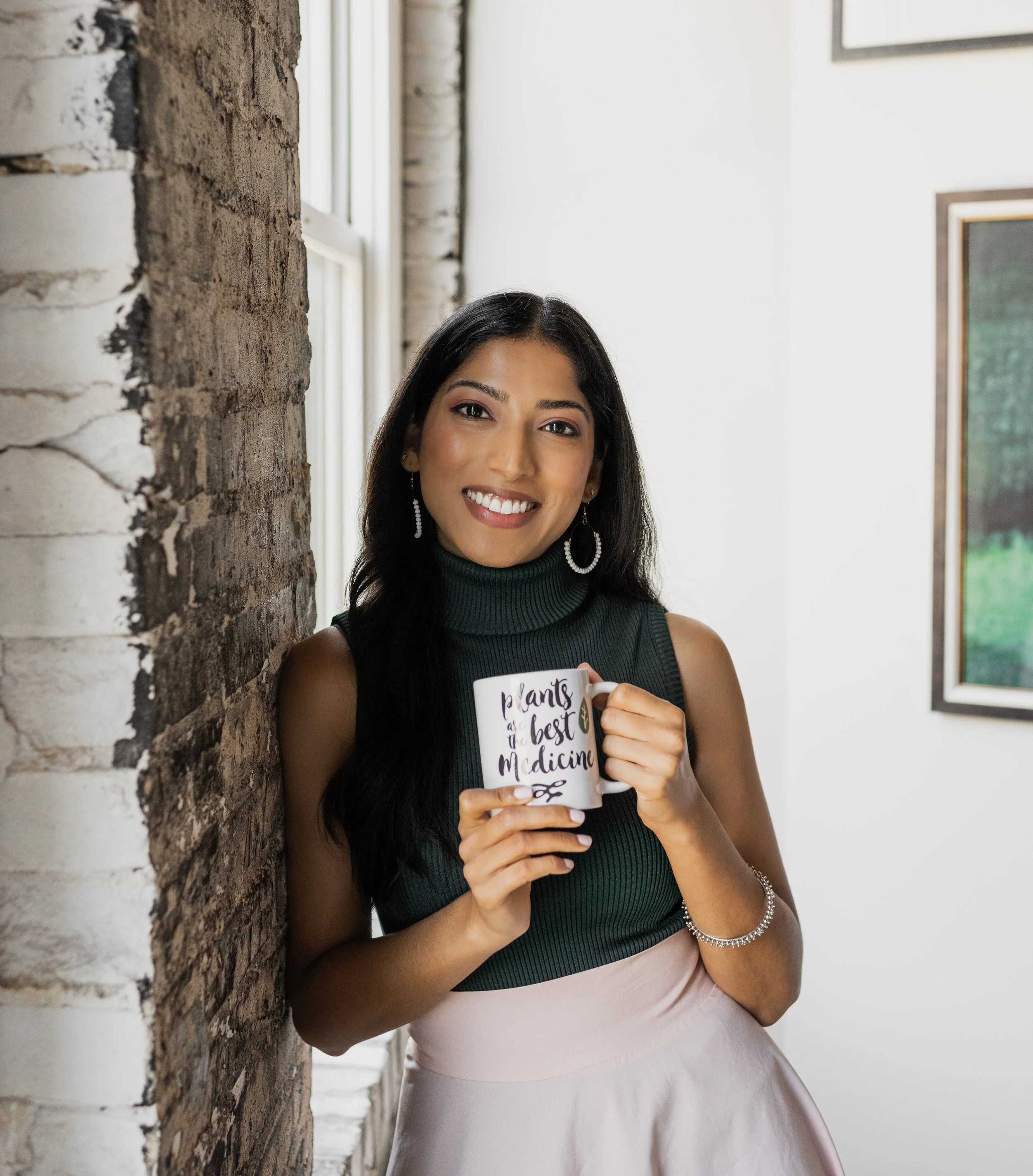 Woman with long black hair smiling and holding a white mug with the text 'plants are the best medicine' in an indoor setting with brick and white walls.