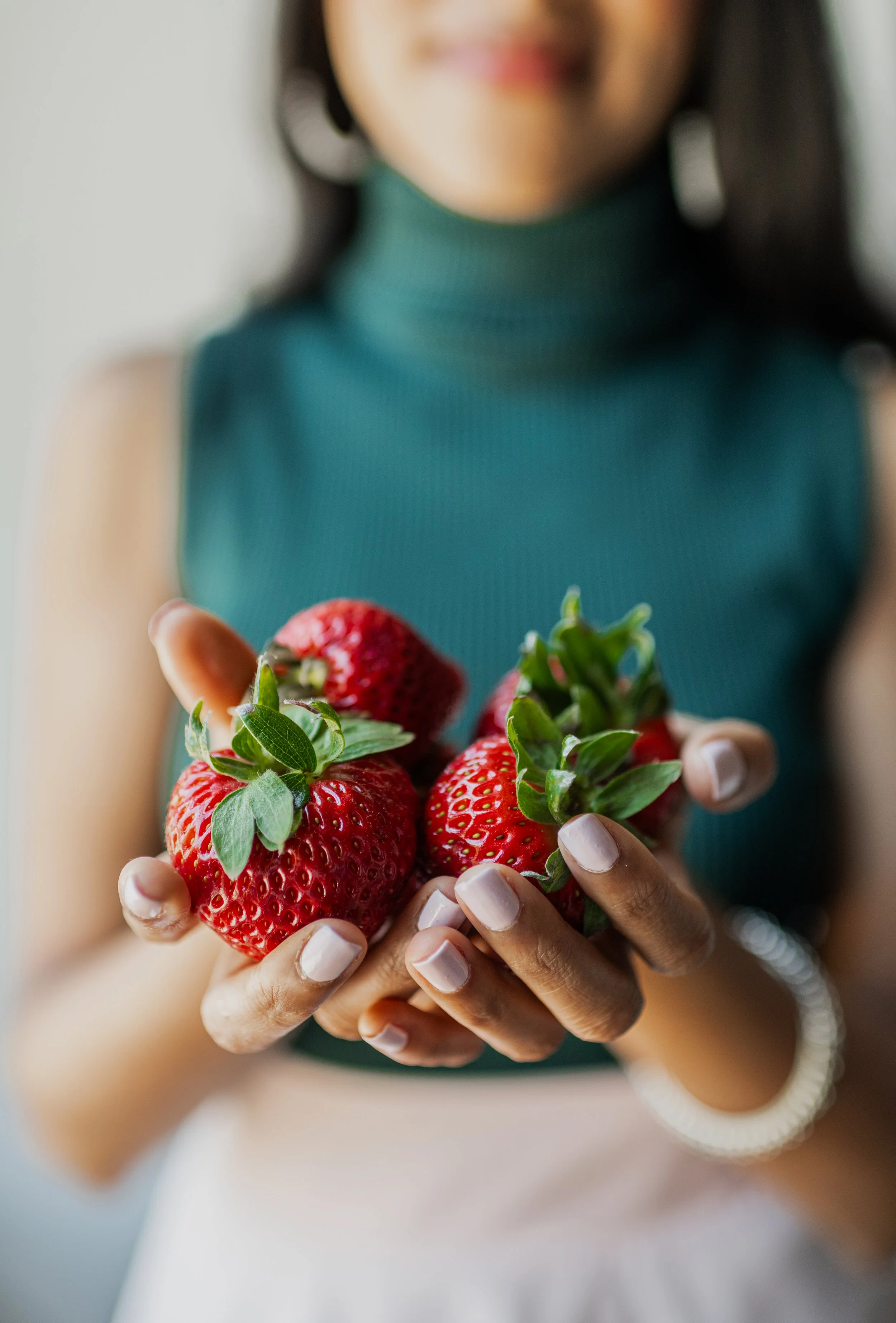 Person holding fresh strawberries with green leaves, wearing a teal sleeveless top.