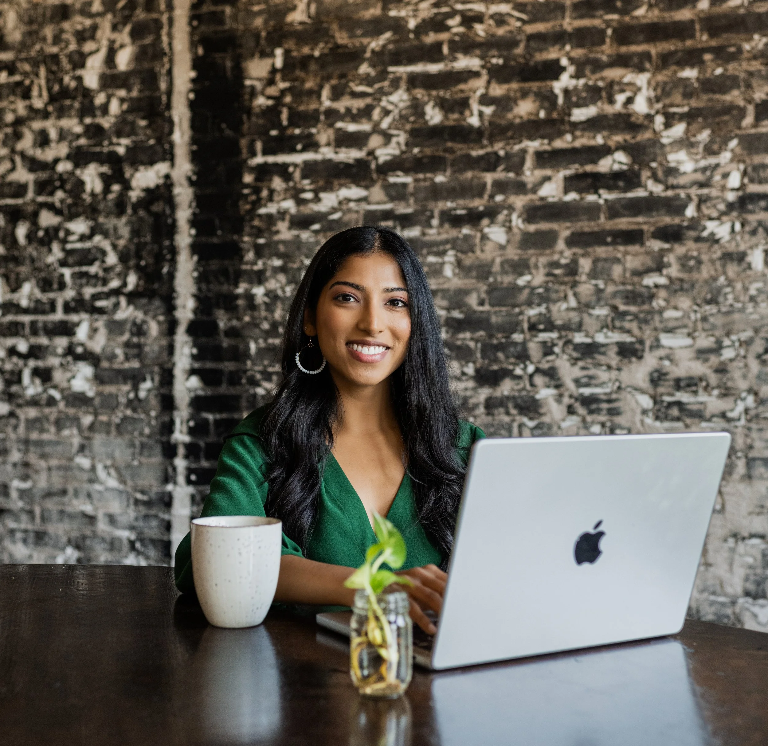 A woman with long black hair and earrings, smiling, sitting at a wooden table with a silver MacBook, a white mug, and a small glass jar with a plant, against a brick wall background.