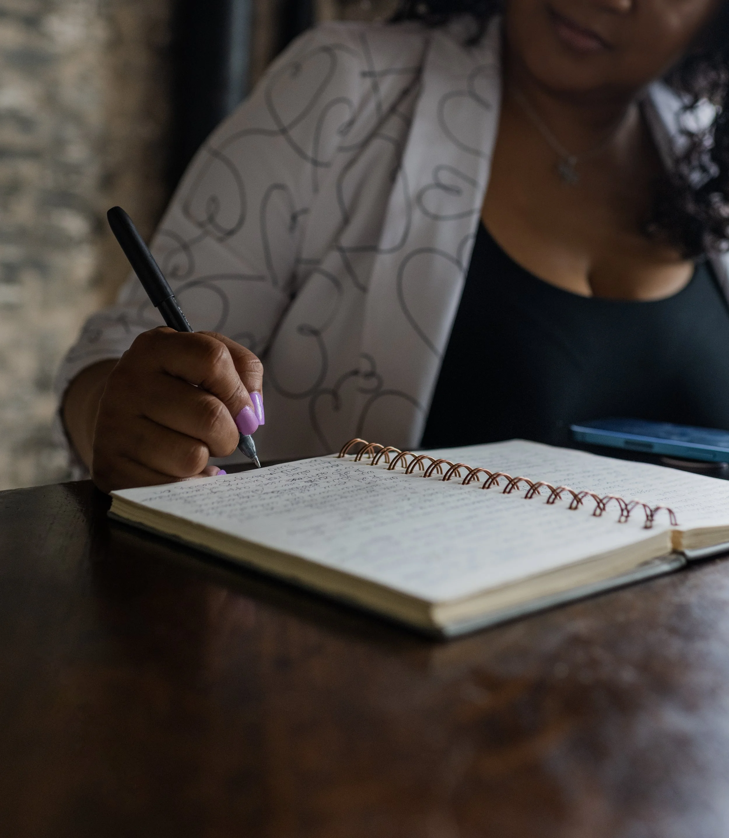 A woman is writing in a spiral notebook at a dark wooden table, with a smartphone nearby.