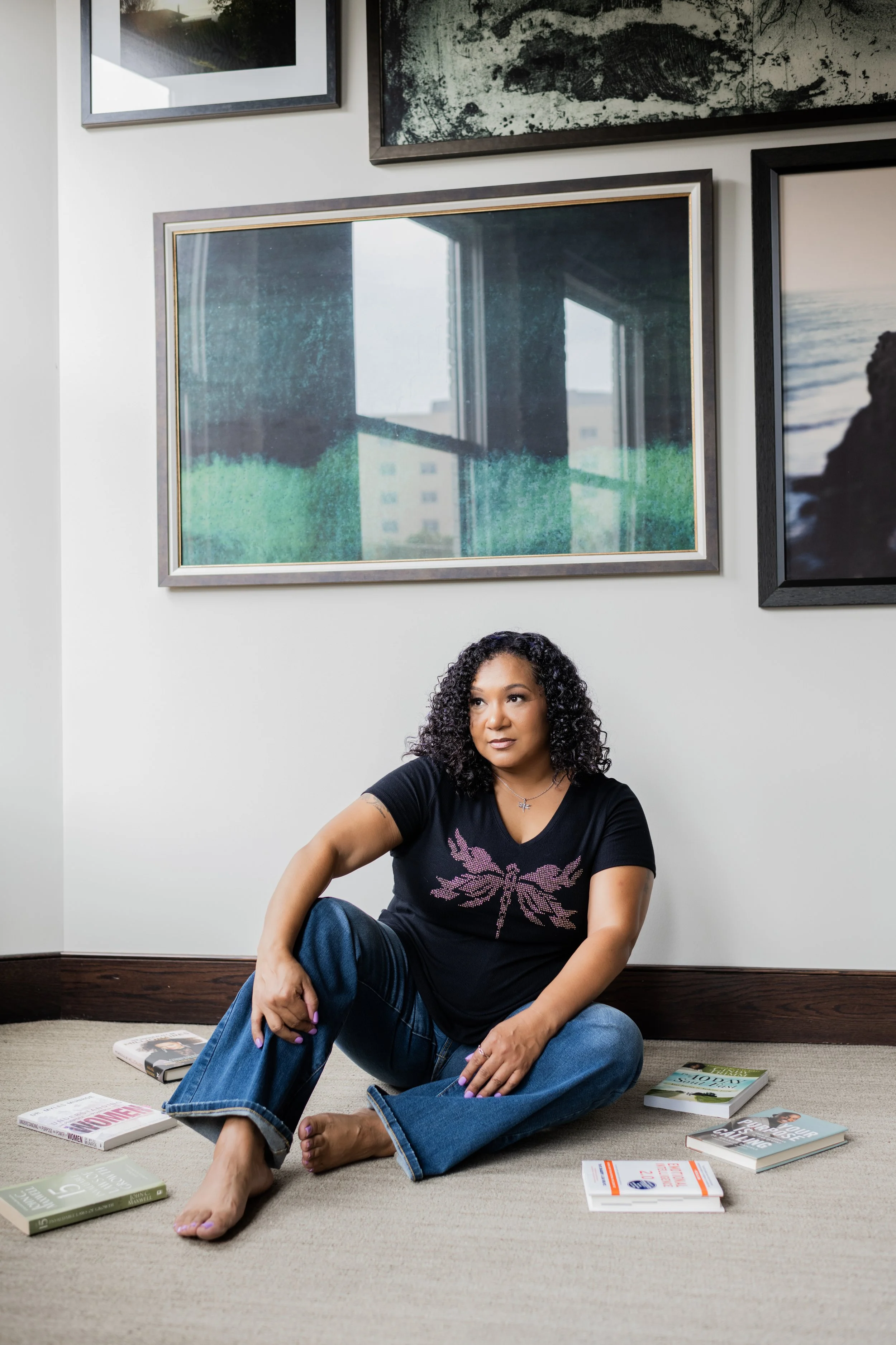 Woman with curly hair sitting on floor with books around her, against a wall with framed photographs.