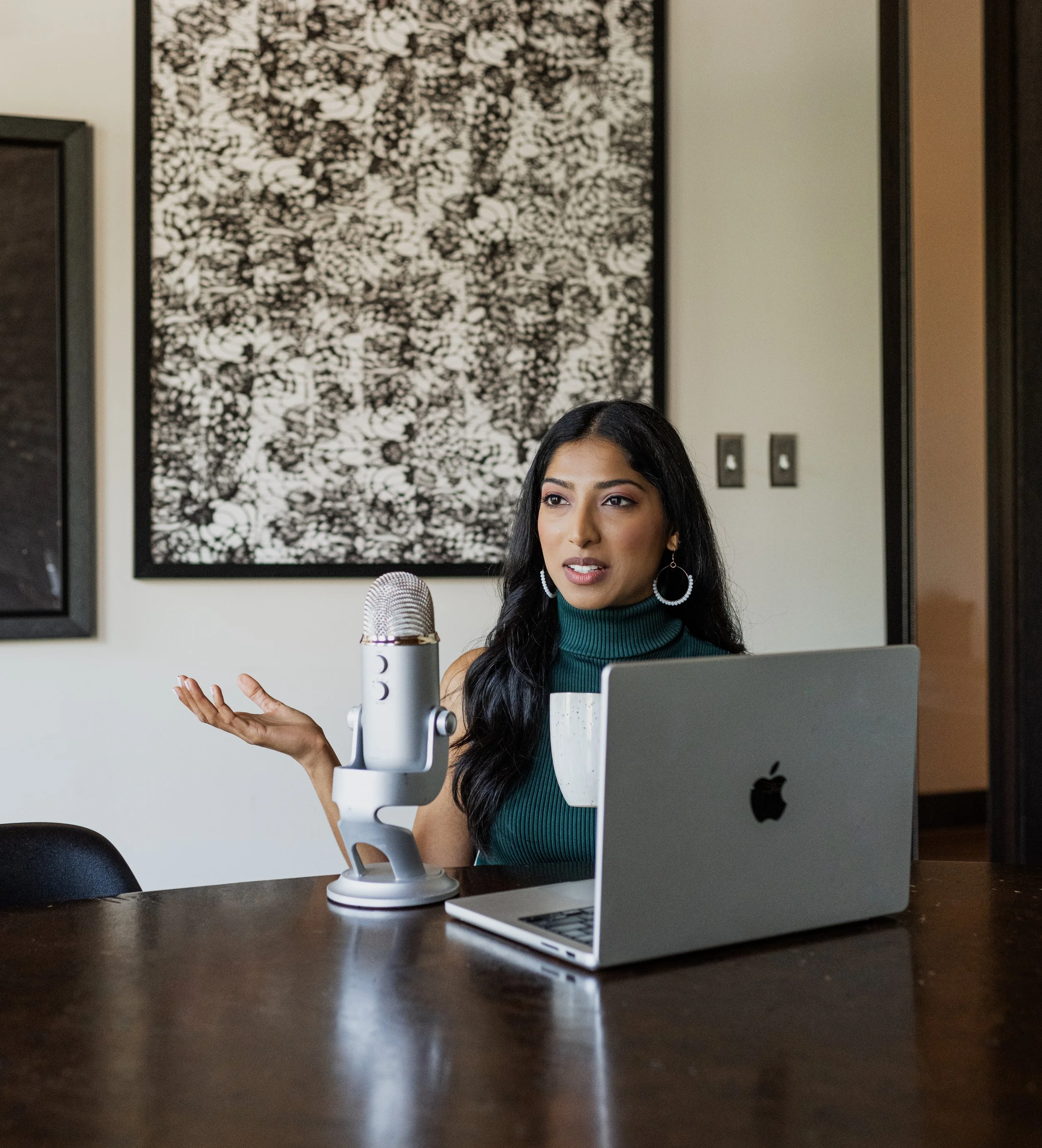 A woman with long black hair sits at a wooden table with a silver MacBook and a white microphone in front of her. She is wearing a dark green sleeveless turtleneck and large hoop earrings, gesturing with her right hand, and appears to be speaking or 