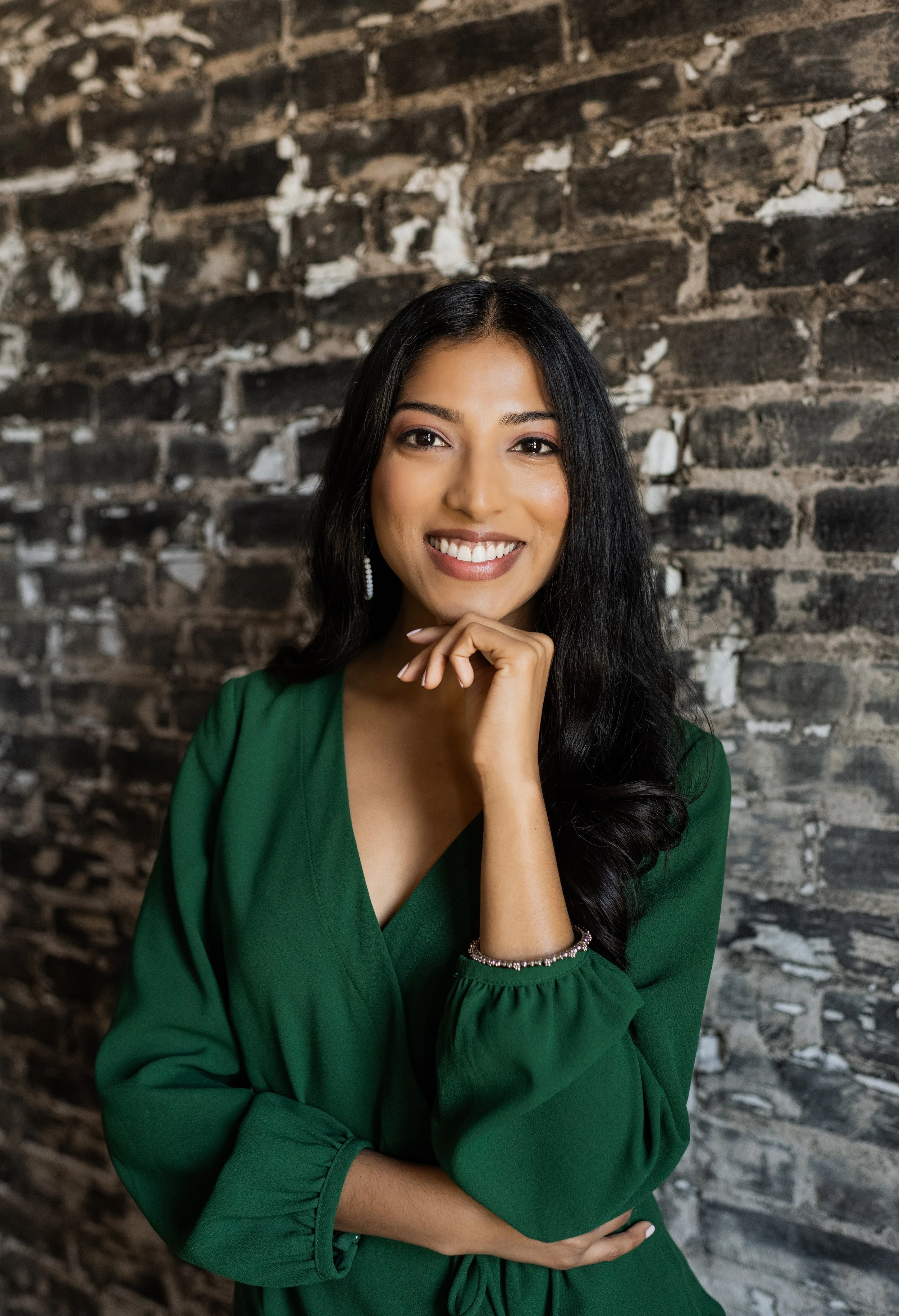 A young woman with long black hair, smiling, wearing a green dress, and standing against a dark brick wall background.