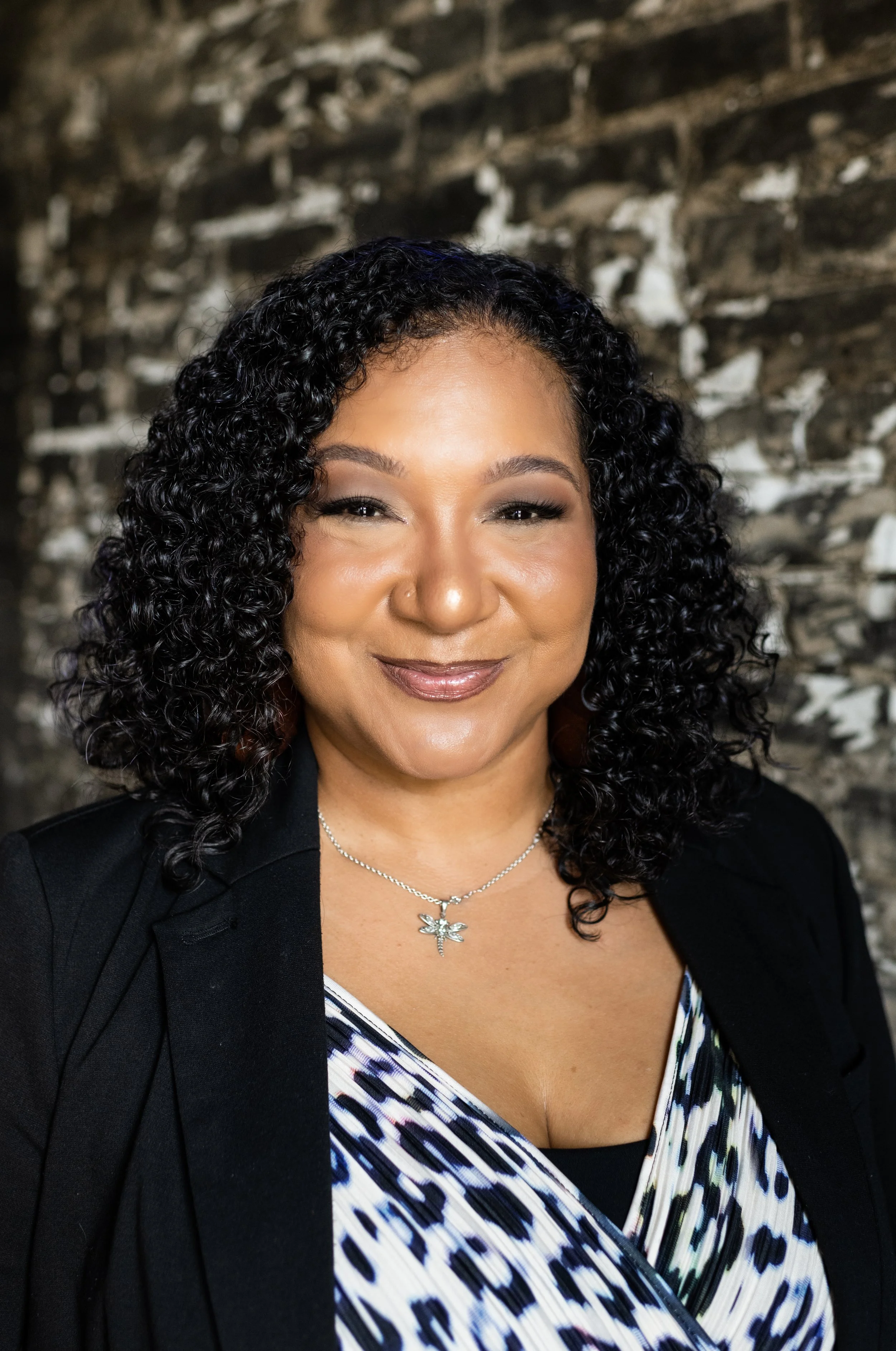 A woman with dark, curly hair smiling, wearing a necklace with a dragonfly pendant, a black blazer, and a patterned top, standing in front of a textured stone wall.