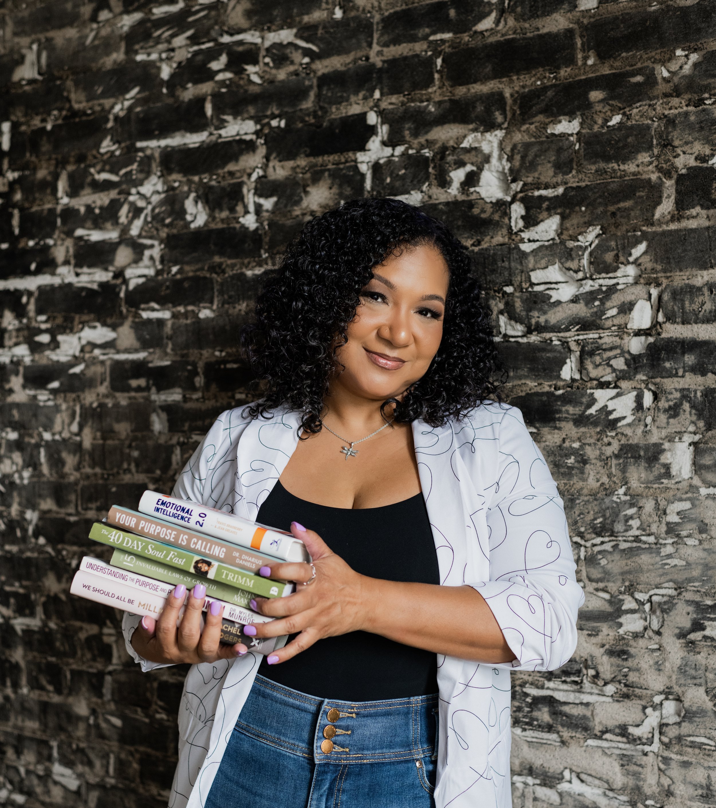 A woman with curly black hair holding a stack of books, standing against a brick wall.