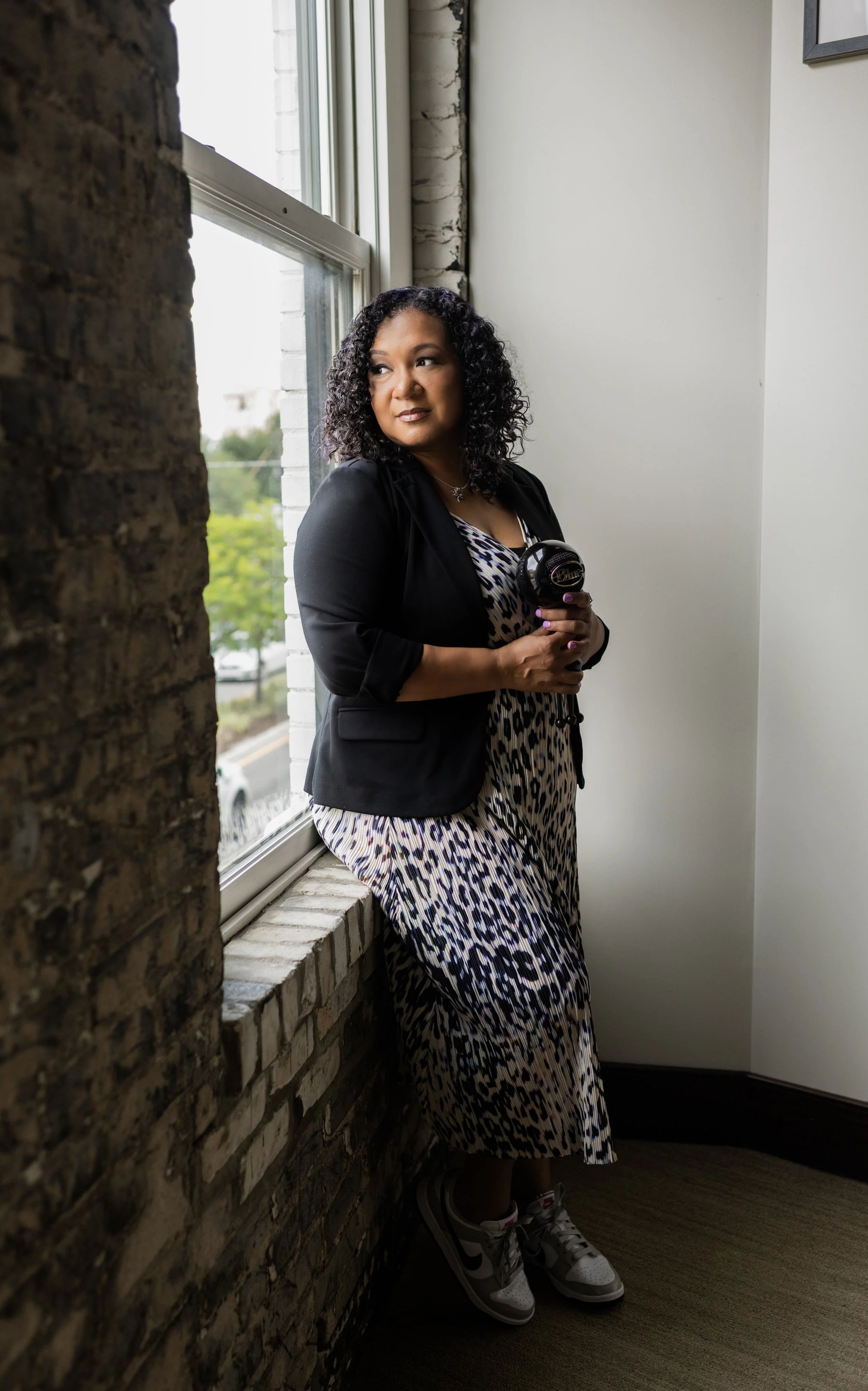 A woman with curly black hair sitting on a window sill, holding a microphone, wearing a leopard print dress, a black blazer, and sneakers, looking out the window.