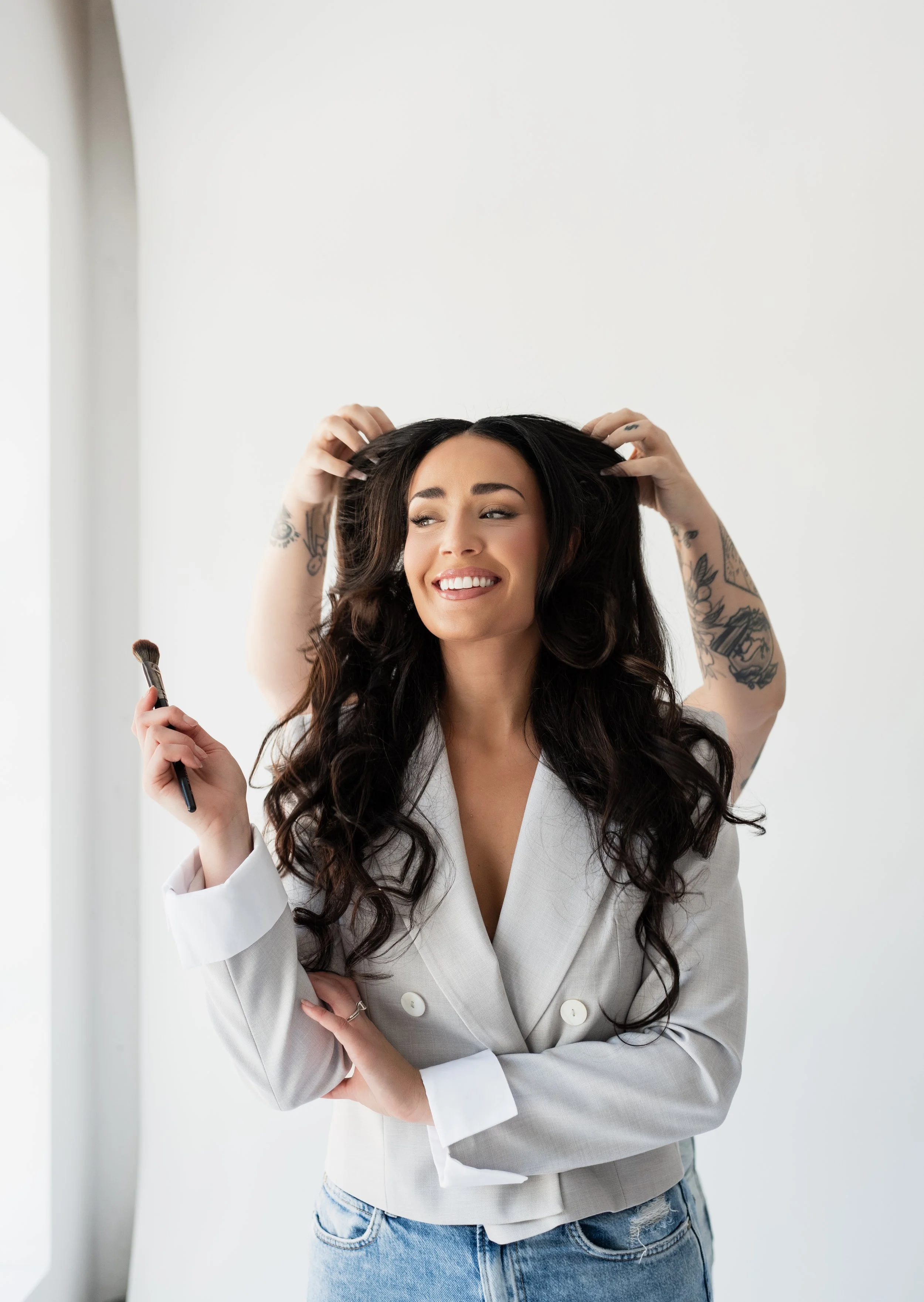 A woman with long dark hair and tattoos, smiling while holding a makeup brush, as a person behind her adjusts her hair.