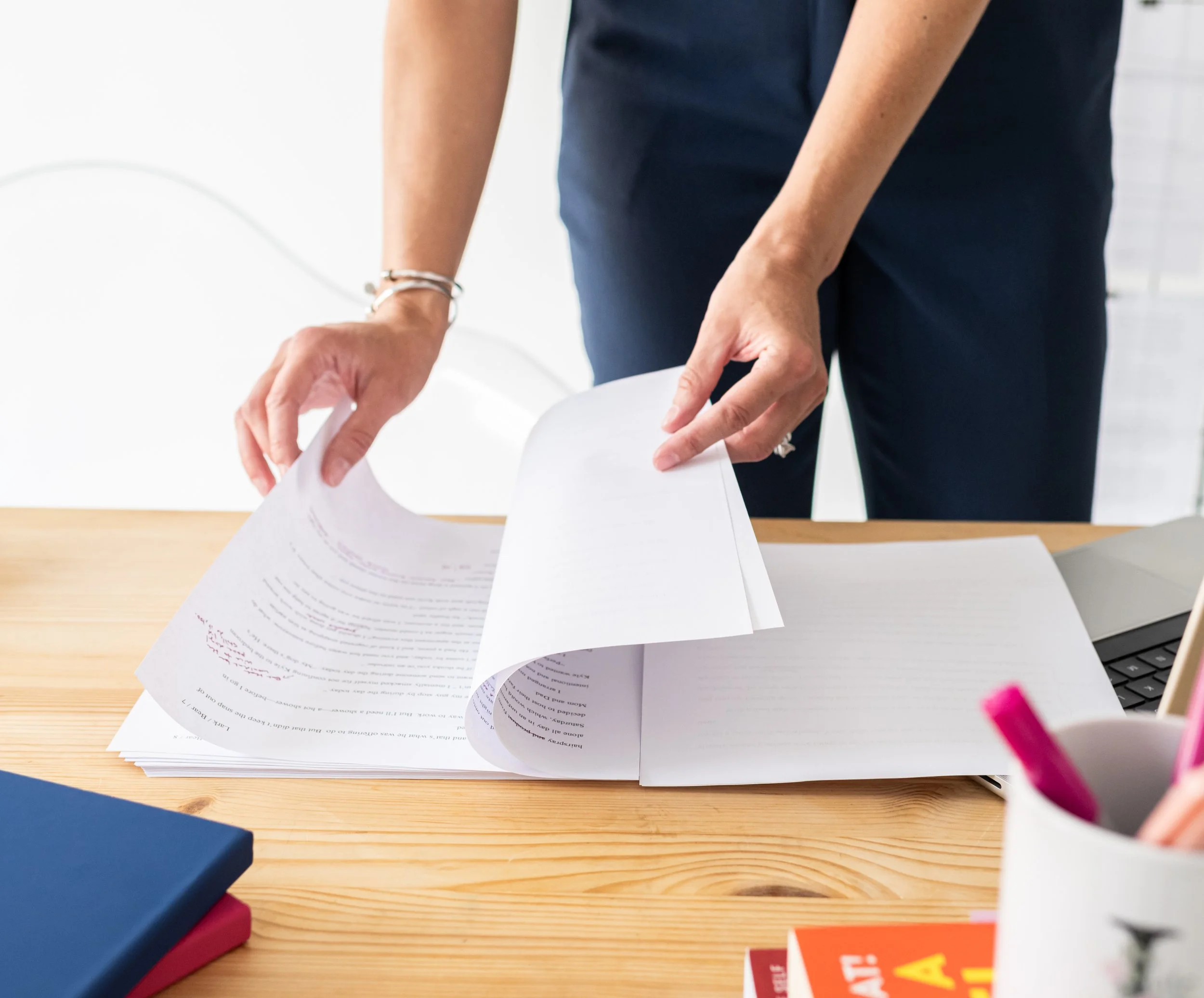 Person with bracelets on wrist flipping through papers on a wooden desk, with a laptop and notebooks nearby.