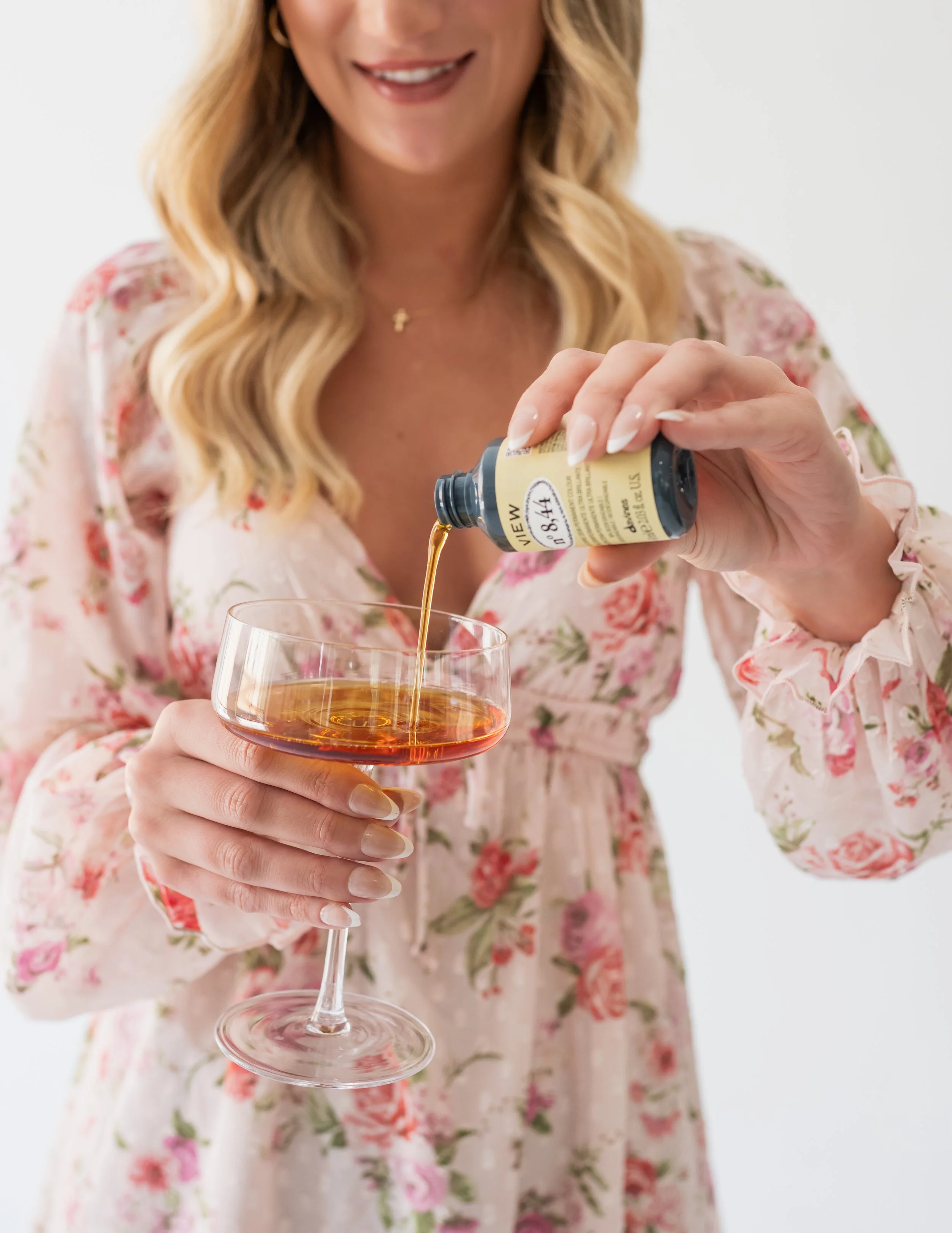 A woman in a floral dress pouring an amber-colored liquid from a bottle into a wine glass.