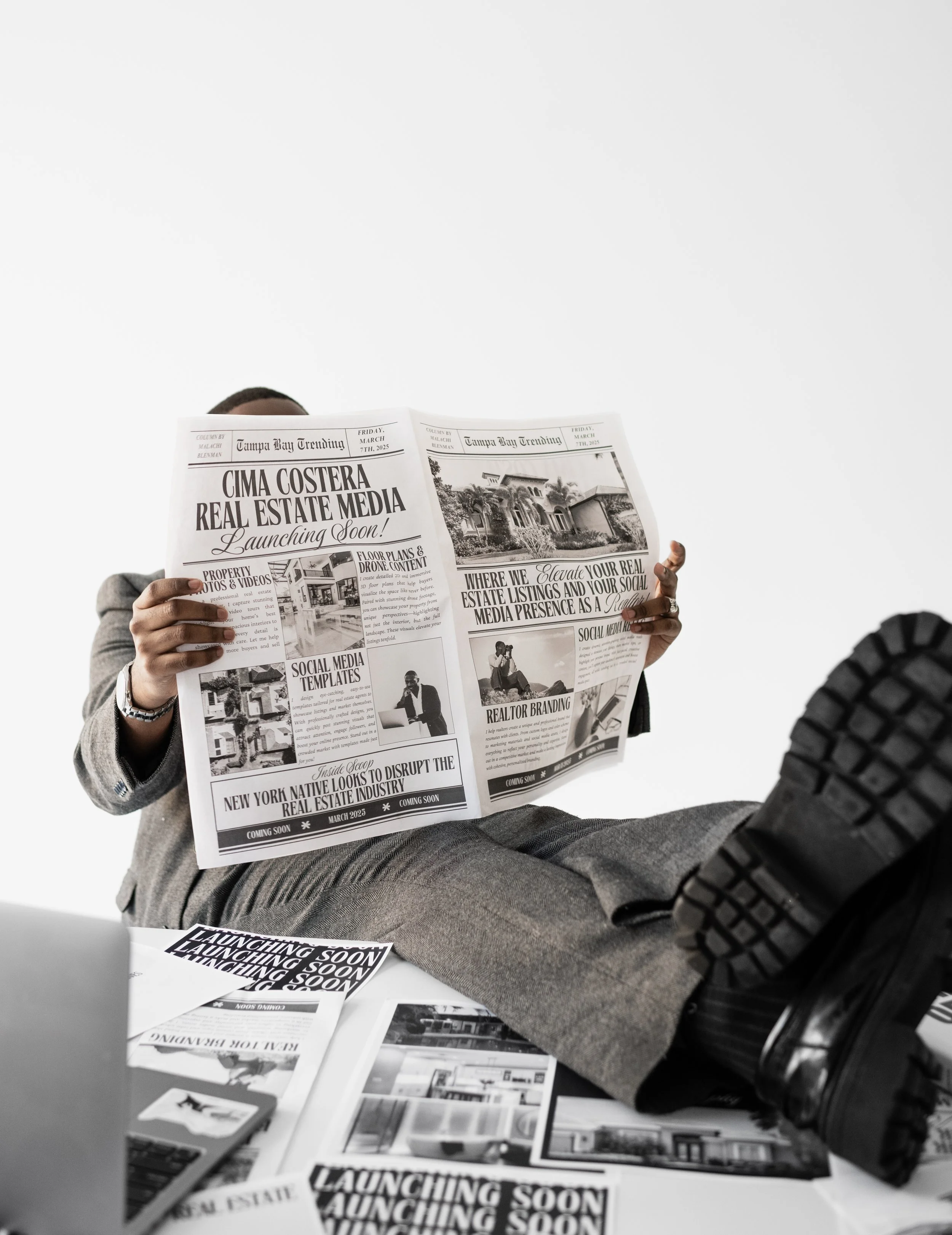 A person sitting at a desk reading a newspaper. The person is dressed in business attire, including gray trousers and a gray coat. The desk has multiple newspapers and documents, and the background is plain white.