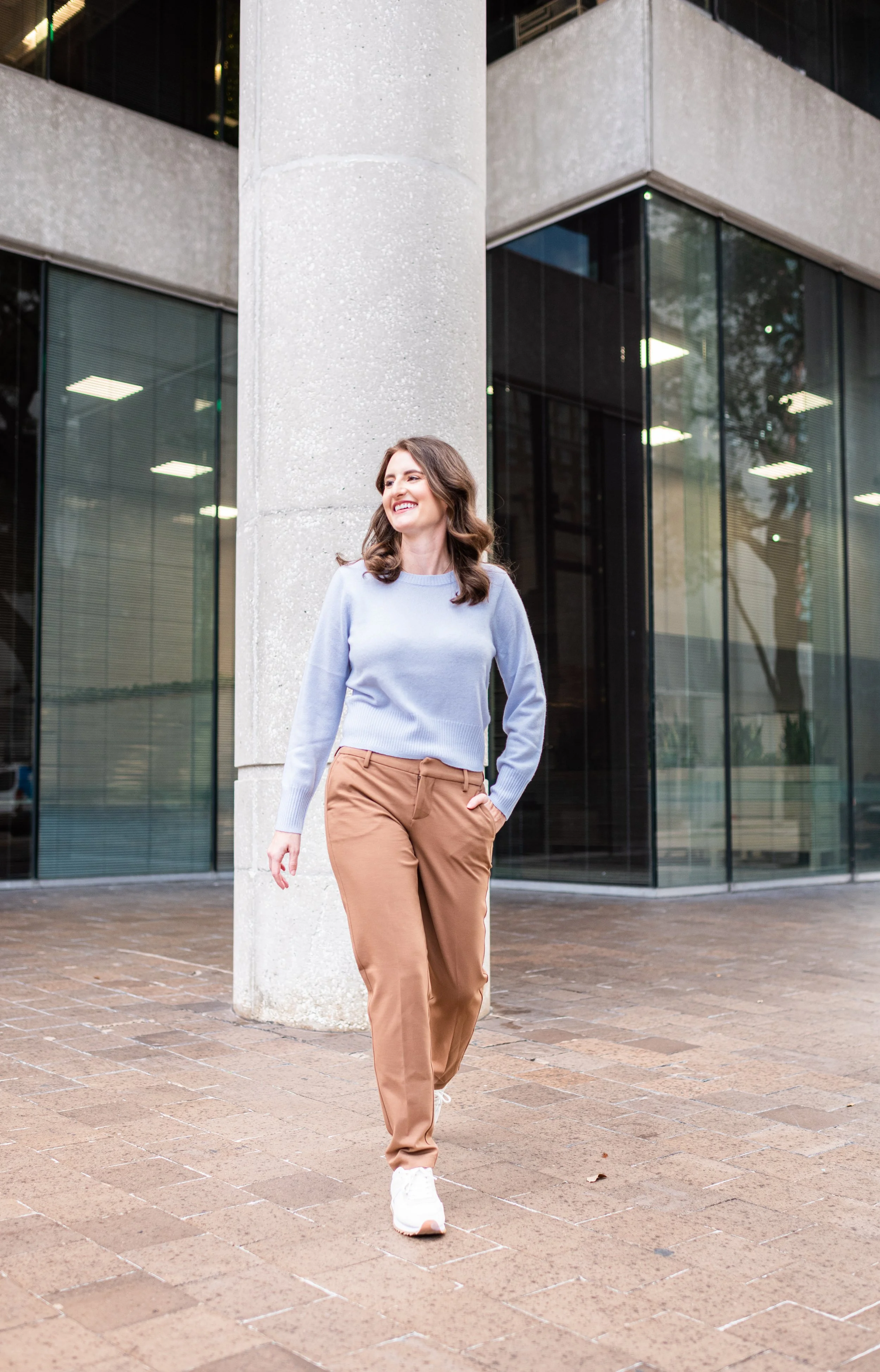 A woman walking outside in front of a modern building with large glass windows, smiling and looking to her right, wearing a light blue sweater, khaki pants, and white sneakers.