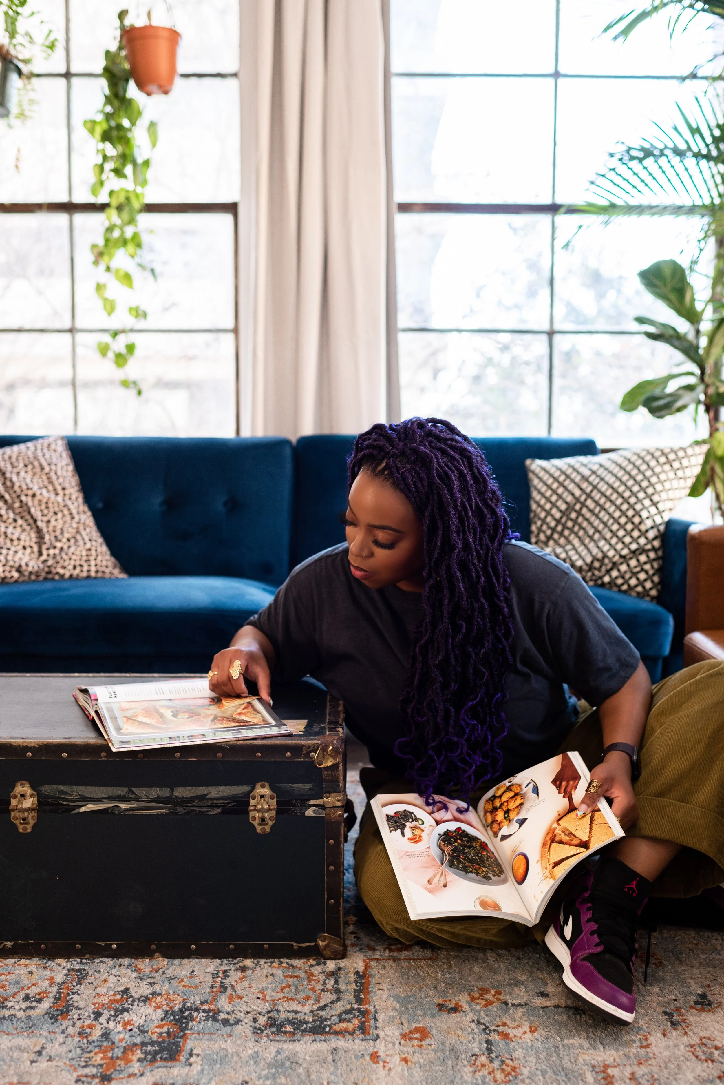 A woman with purple dreadlocks reading a magazine while sitting on the floor in a living room with large windows, a blue sofa, patterned pillows, and plants.