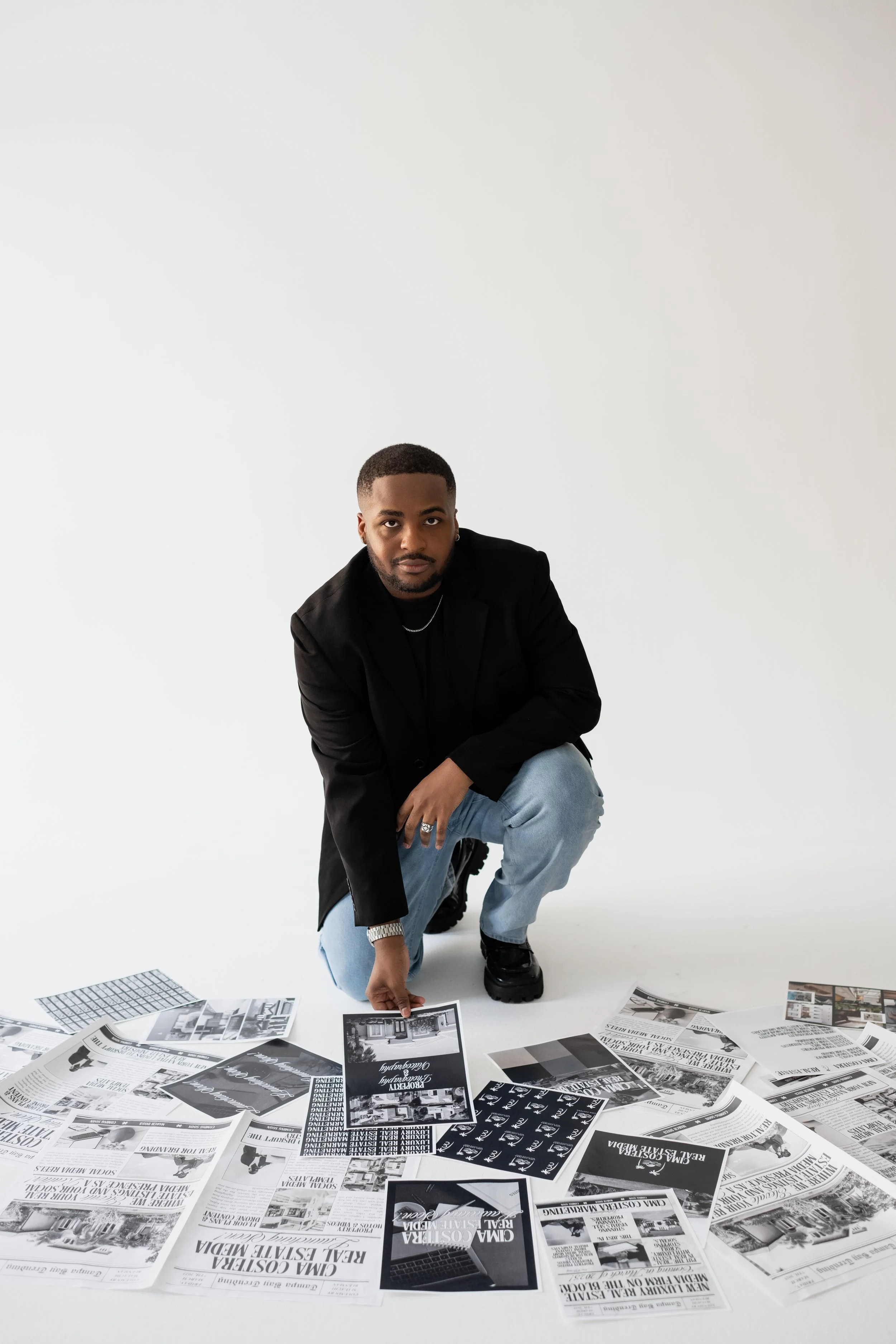 A man in a black jacket and light blue jeans crouching on the floor, surrounded by newspapers and photographs, holding a black and white photo.