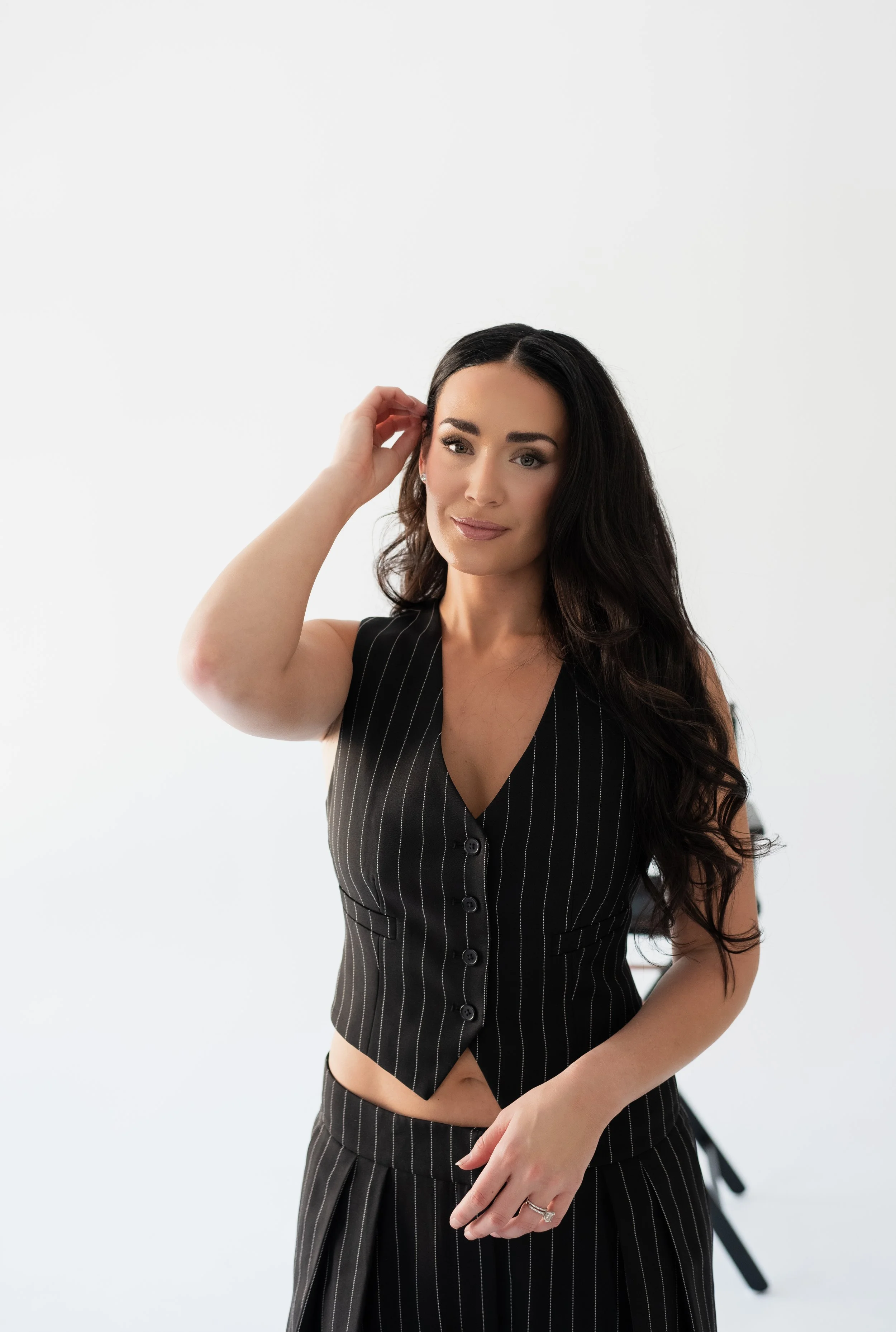 A woman with long dark hair, wearing a black pinstripe vest and matching skirt, posing against a plain white background.