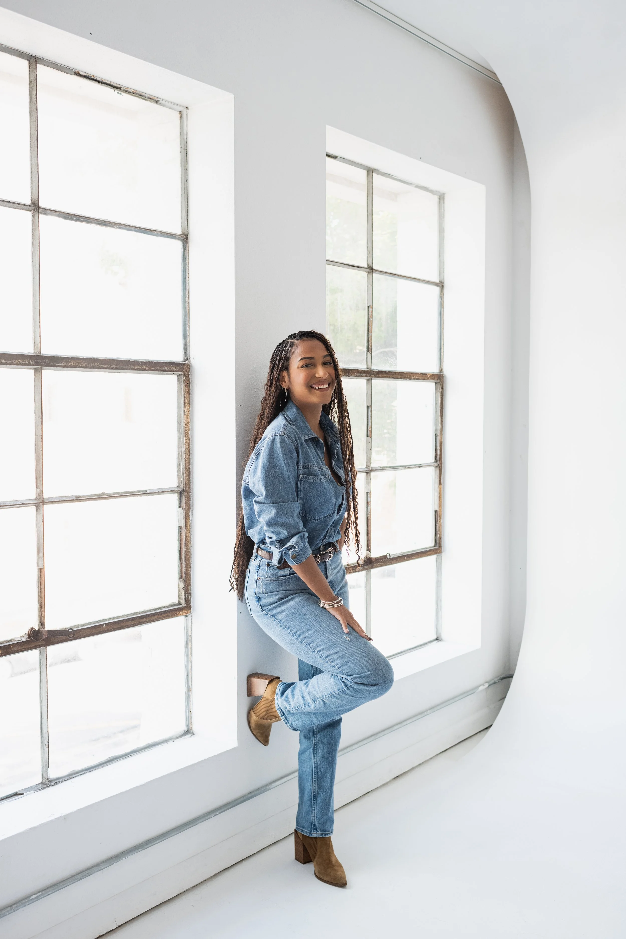 Smiling young woman in denim outfit posing indoors near large windows with white walls.