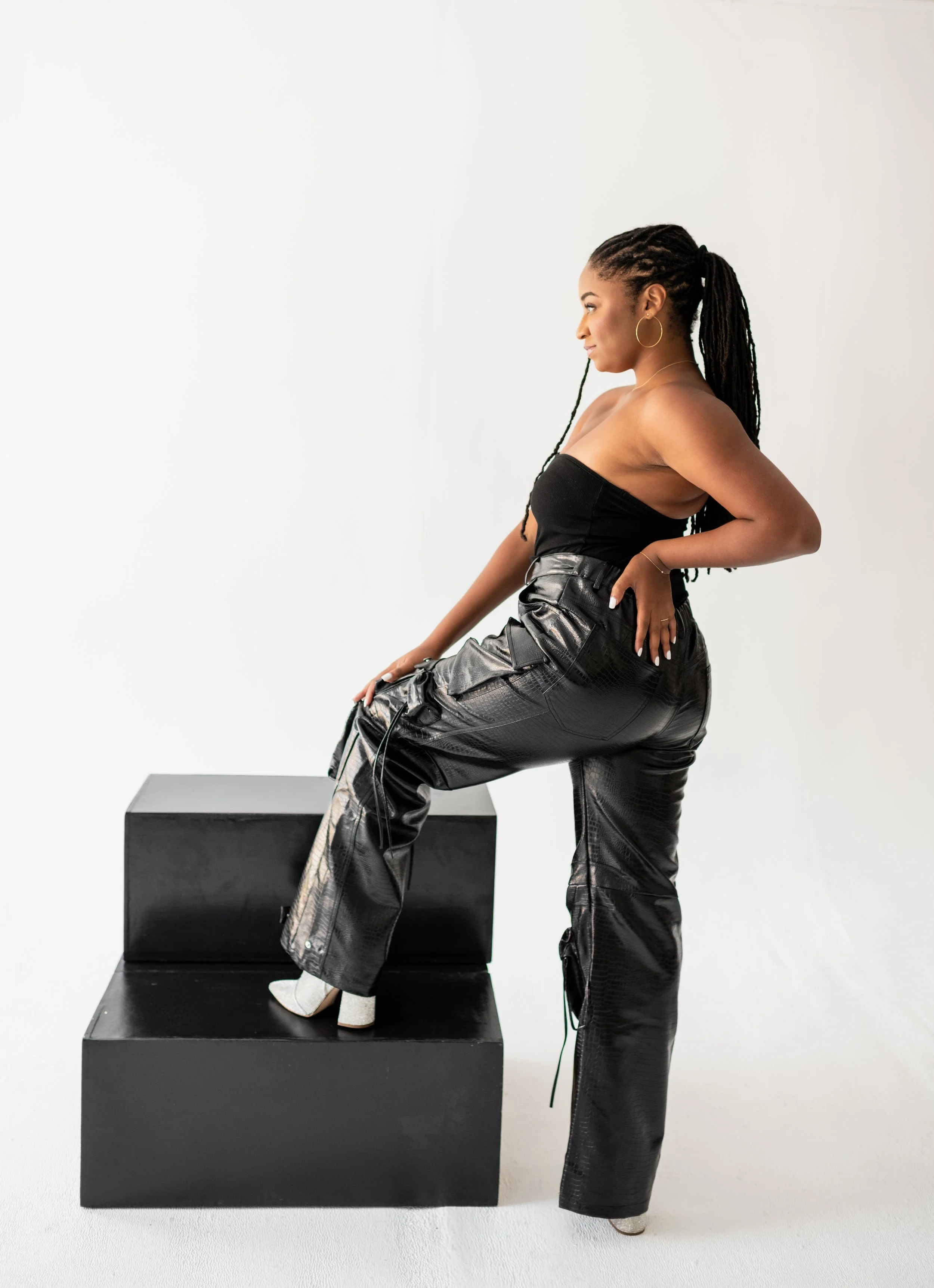 A woman with dreadlocks wearing a black strapless top, black leather pants, and white high-heeled boots posing with one foot on black steps against a white background.