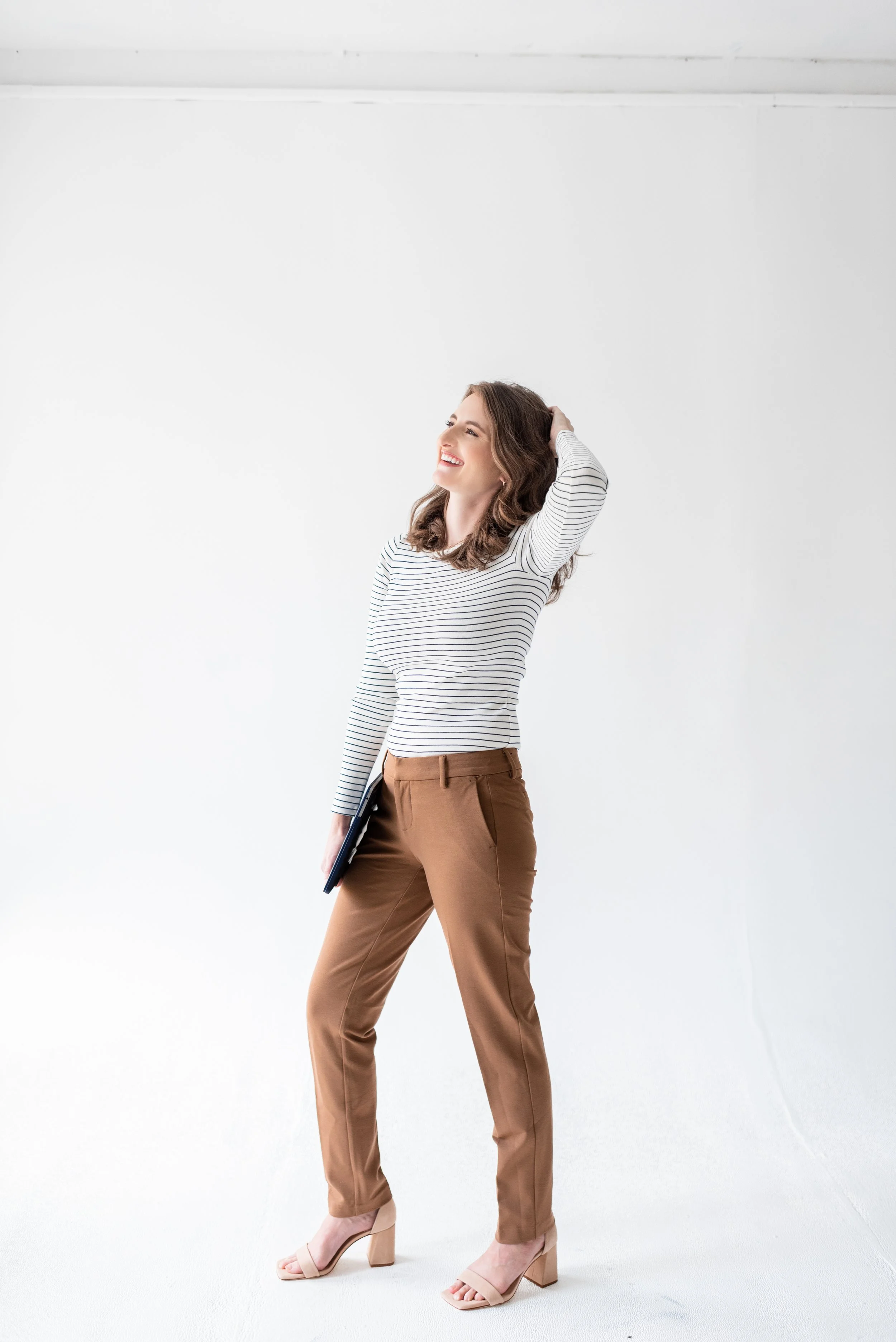 A woman standing against a white background, smiling, wearing a striped long sleeve top, brown pants, and beige high heels, holding a closed laptop in her left hand.