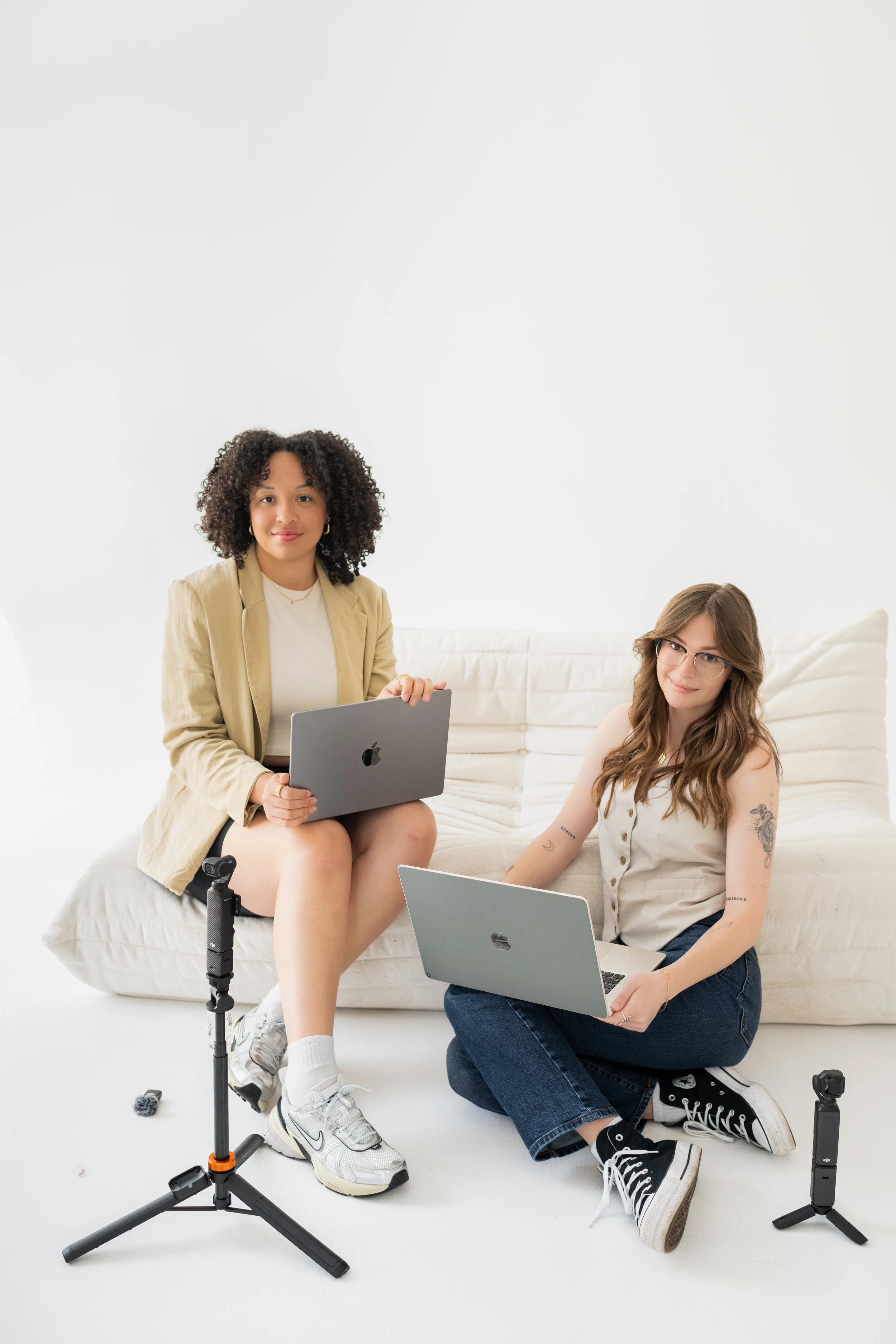 Two women sitting on a white couch with laptops, with tripods in front, one standing and one sitting, in a minimalistic white room.