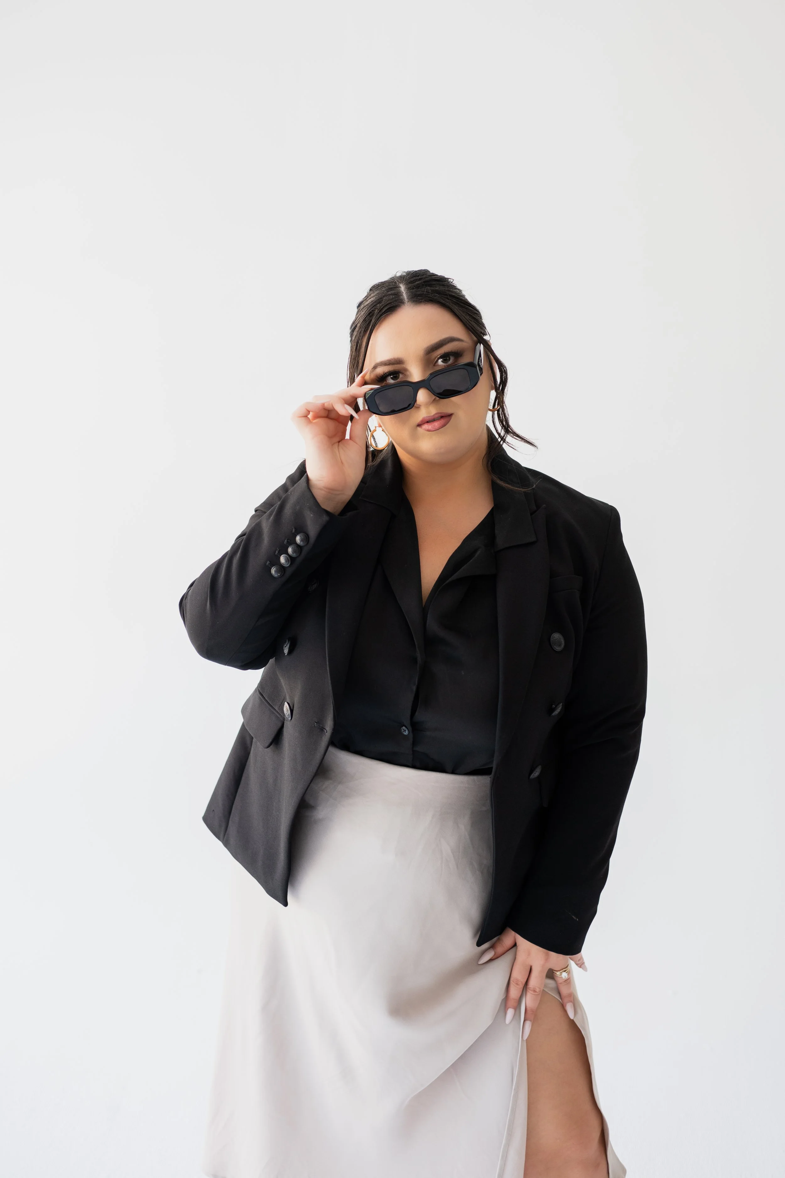 A woman with dark hair and makeup wearing a black blazer and shirt, a light gray skirt with a slit, and sunglasses, posing against a plain white background.